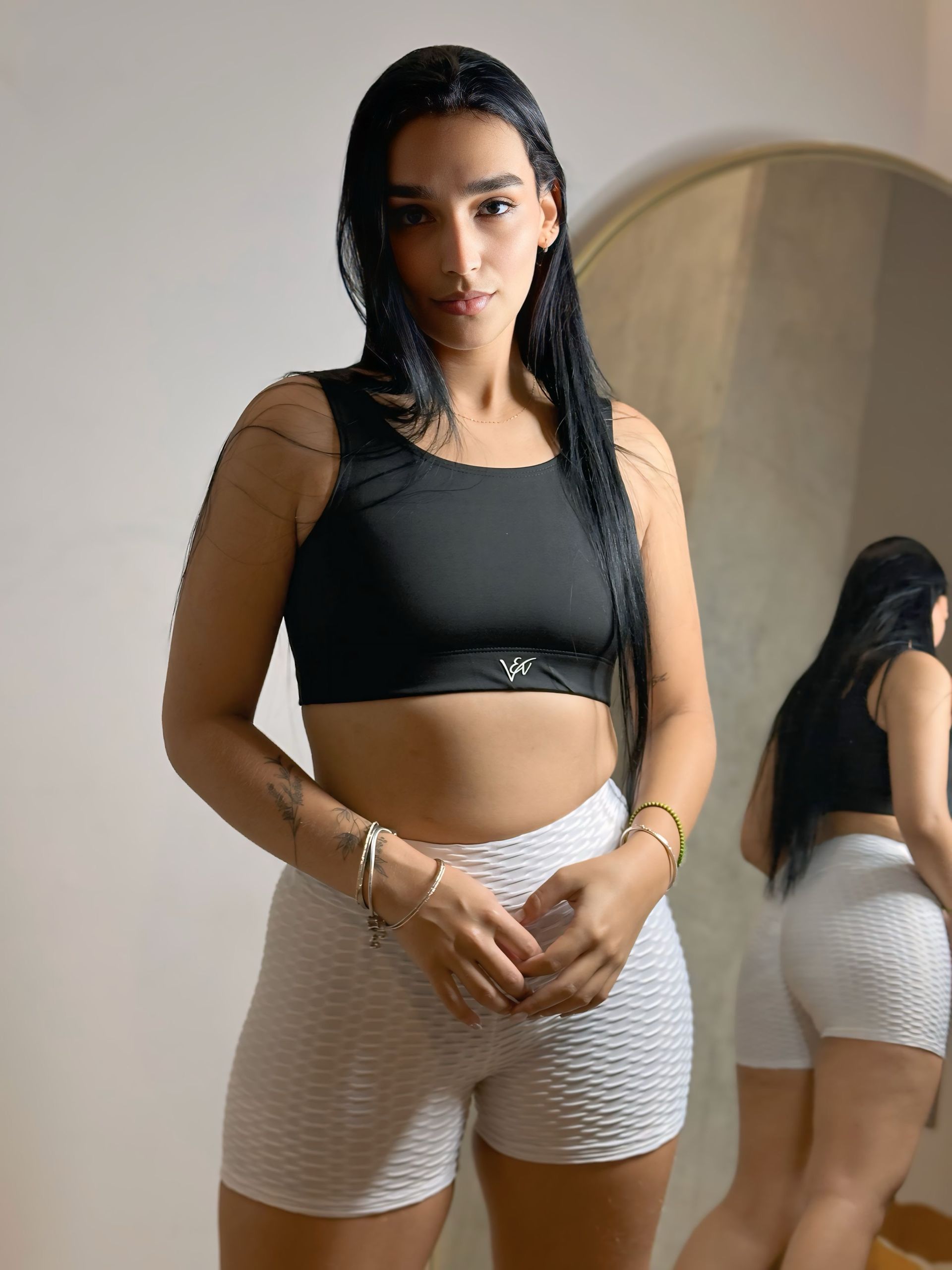 A woman in a black crop top and white shorts is standing in front of a mirror.