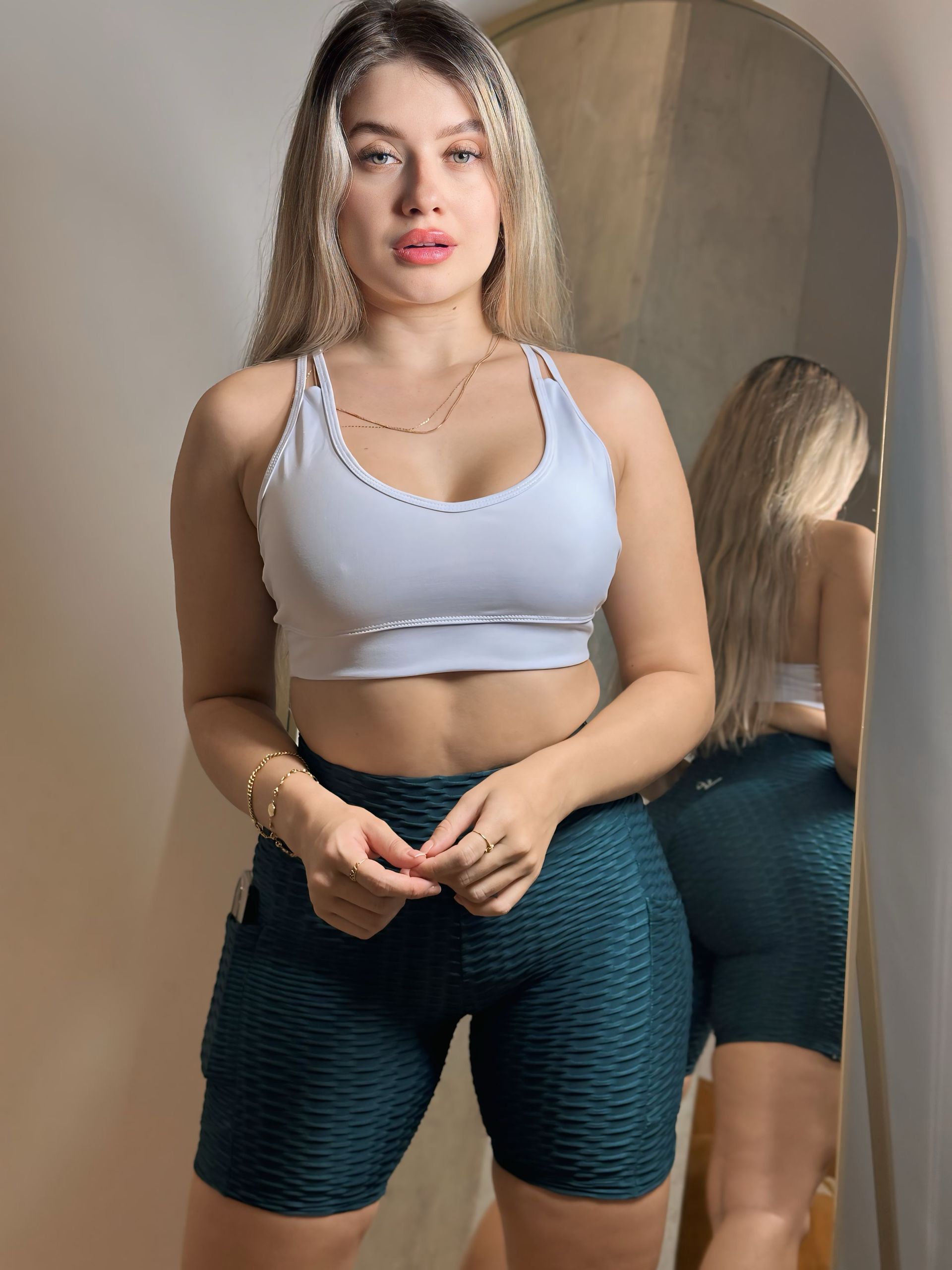A woman in a white crop top and green shorts is standing in front of a mirror.