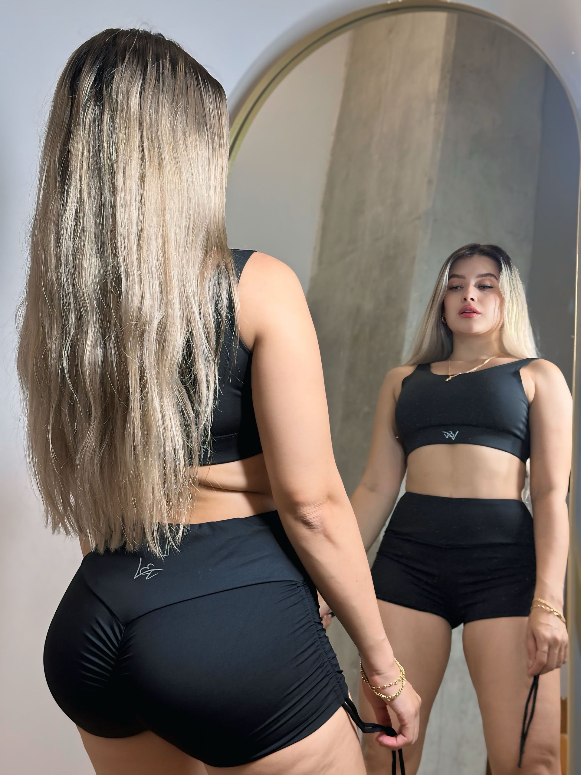 A woman in a black top and shorts is standing in front of a mirror.