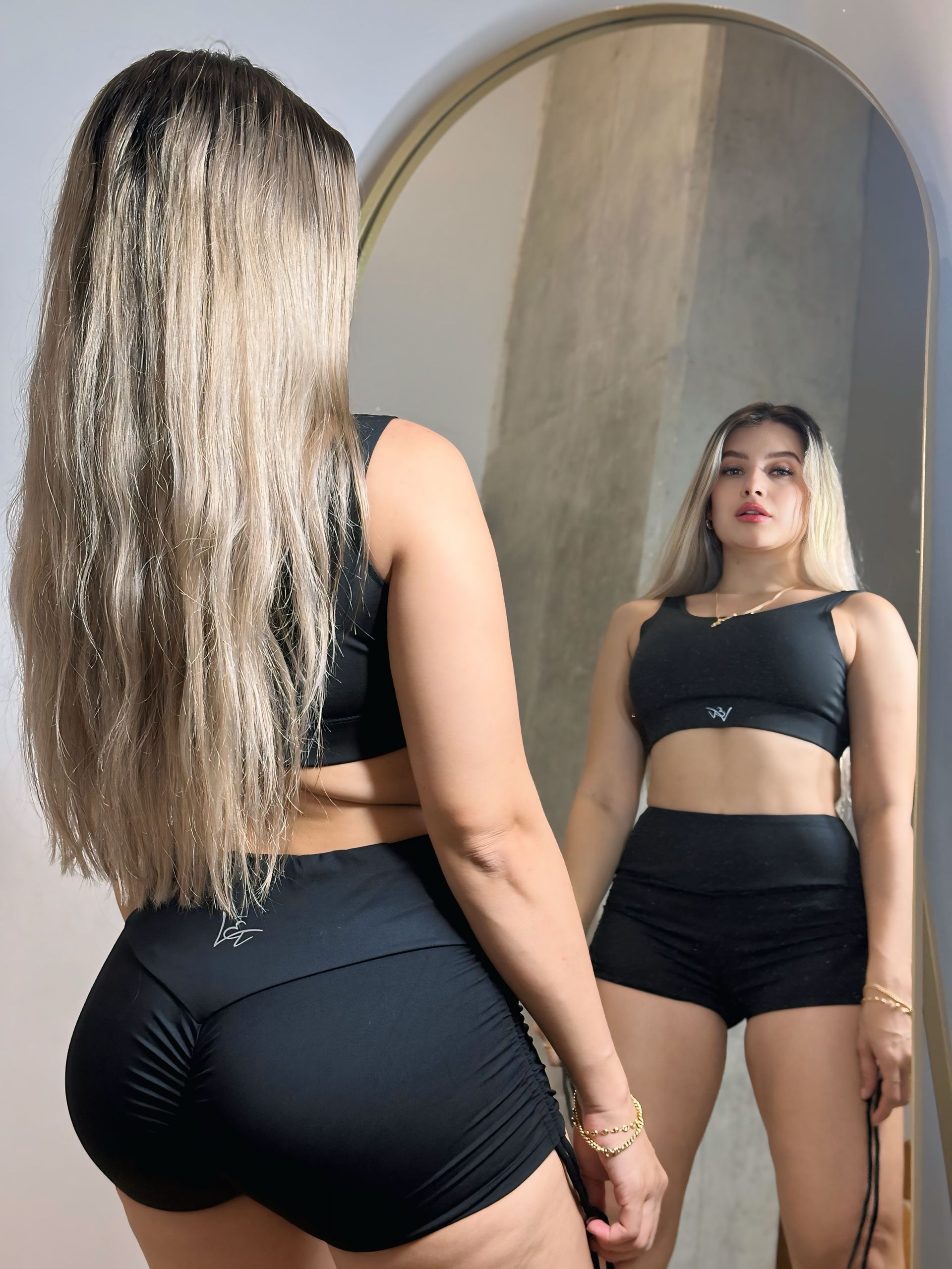 A woman in a black top and shorts is standing in front of a mirror.
