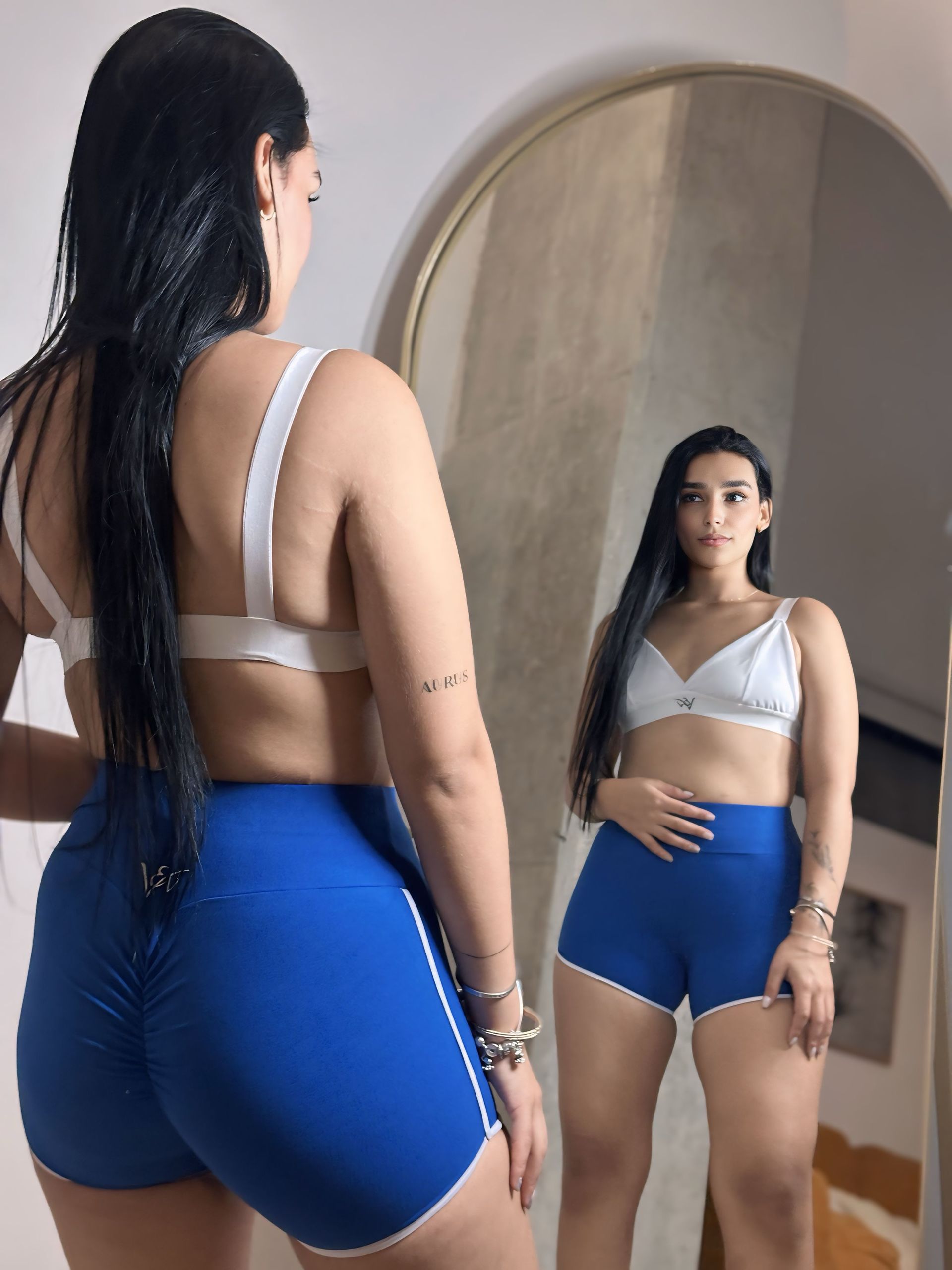 A woman is standing in front of a mirror wearing blue shorts and a white bra.