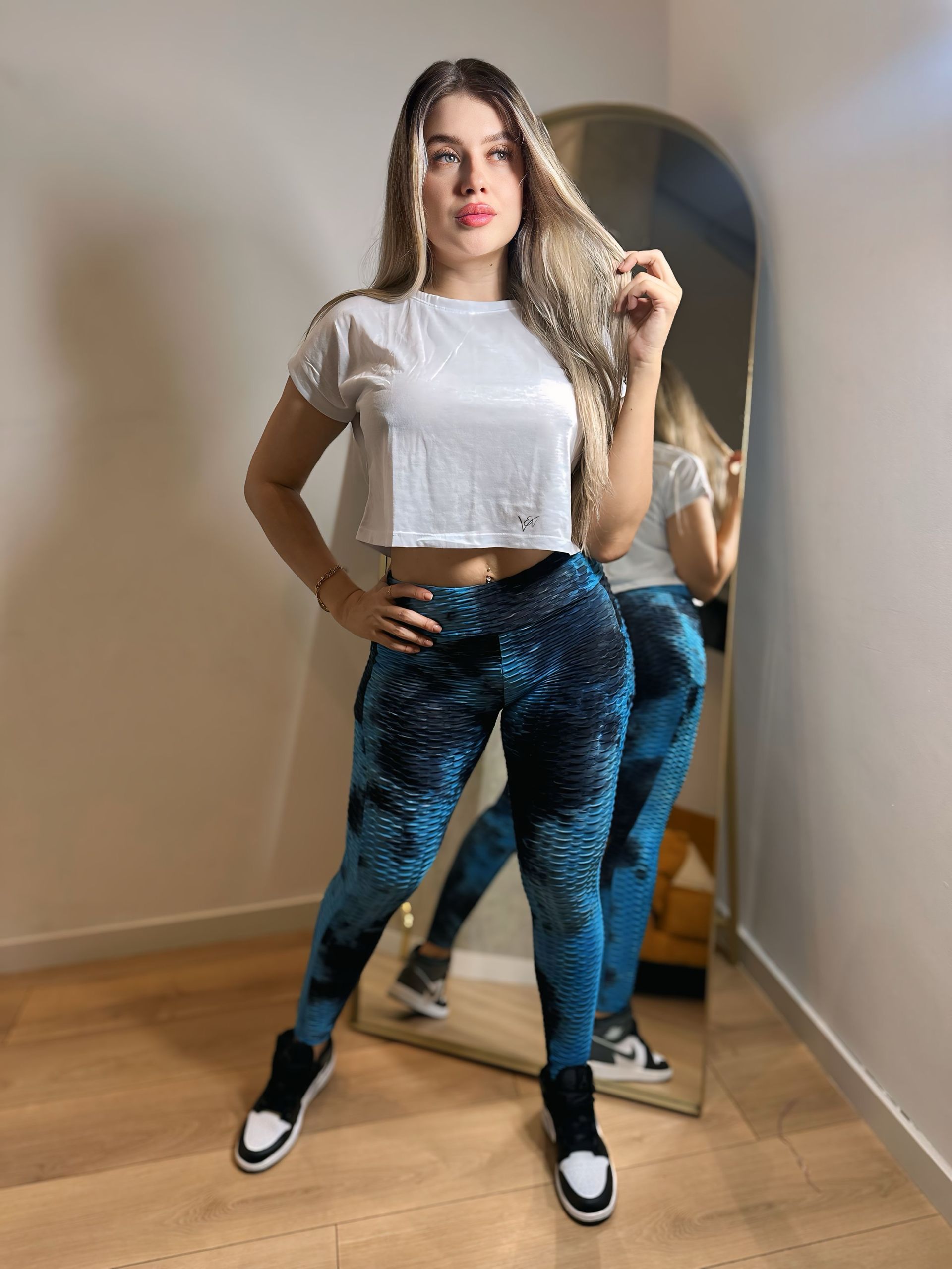 A woman is standing in front of a mirror wearing a white crop top and blue leggings.