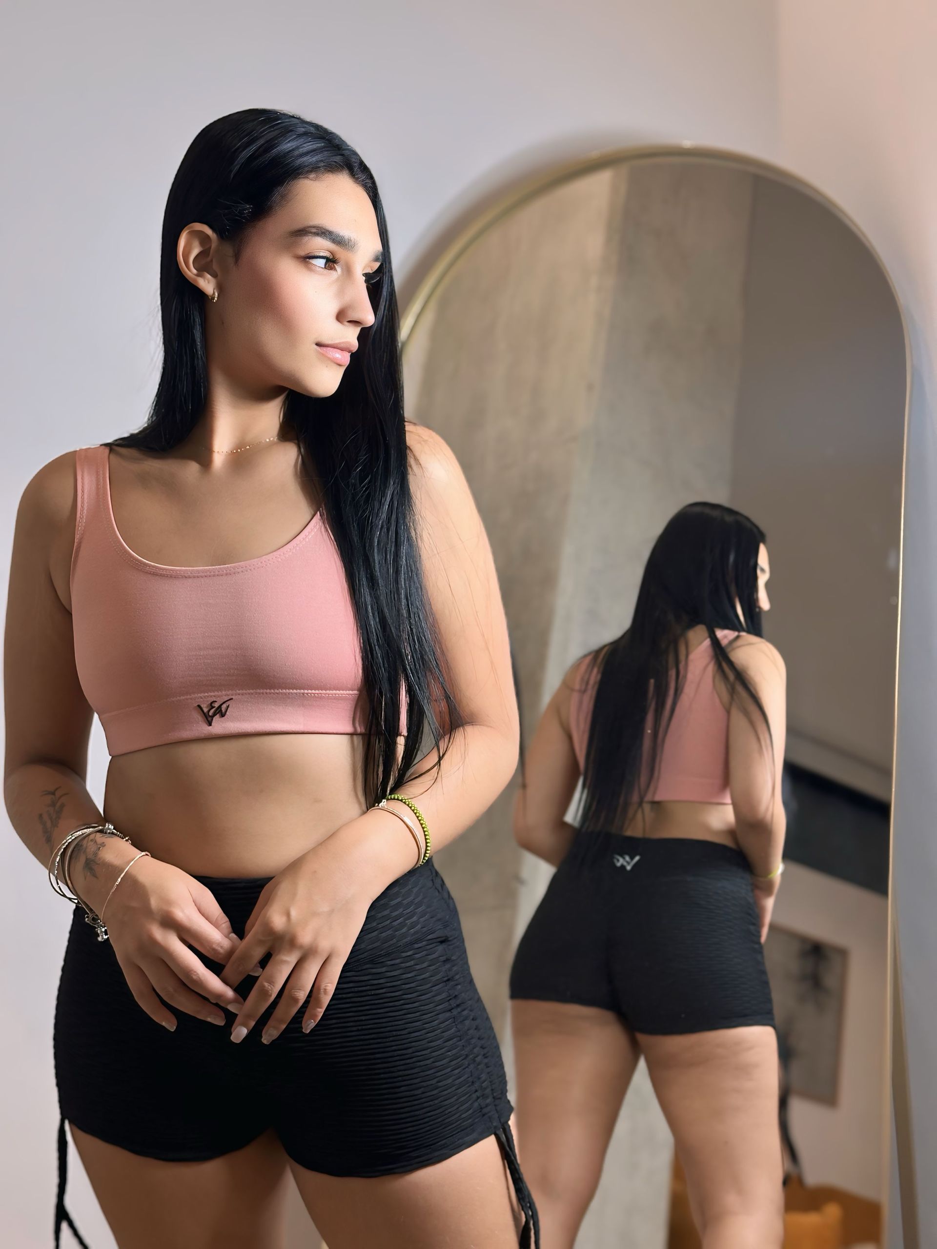 A woman in a pink crop top and black shorts is standing in front of a mirror.