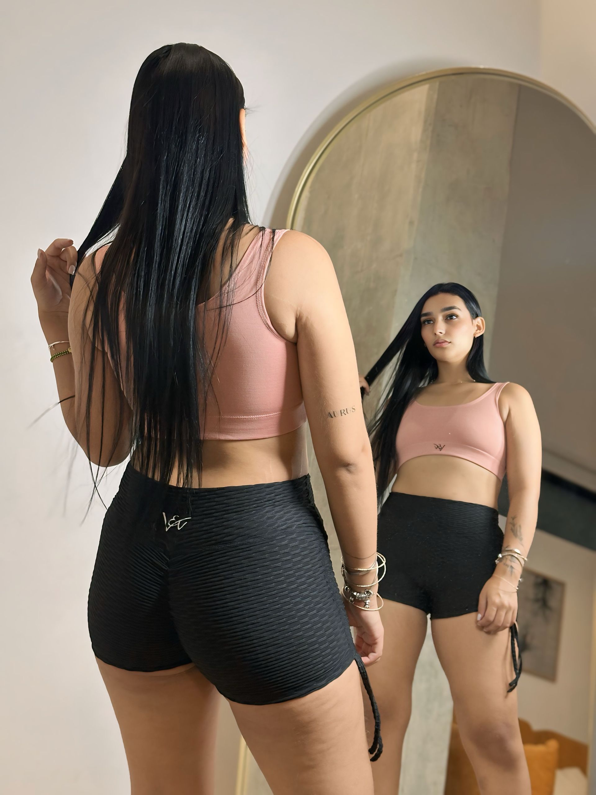 A woman in a pink top and black shorts is standing in front of a mirror.