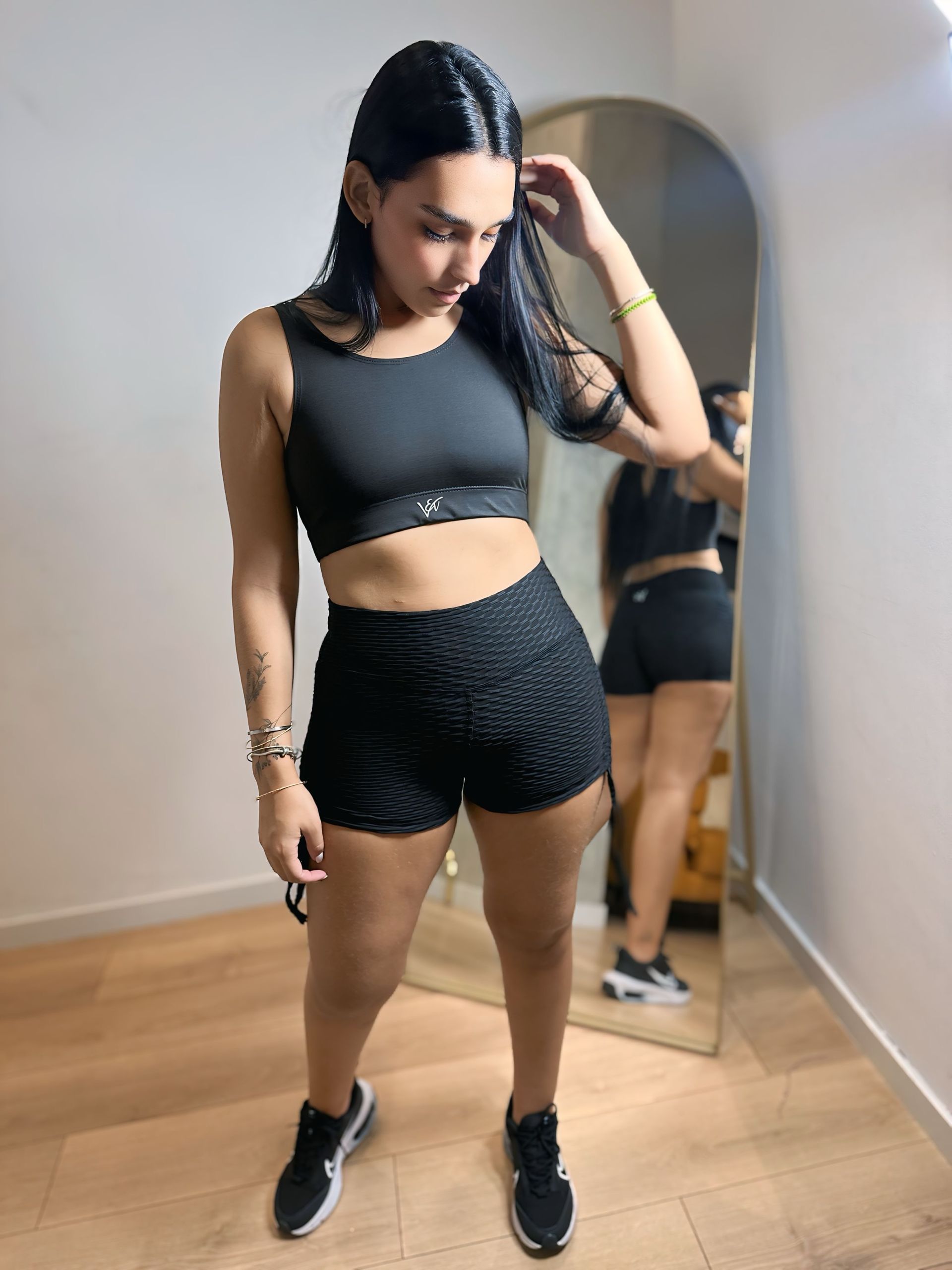 A woman in a black crop top and shorts is standing in front of a mirror.