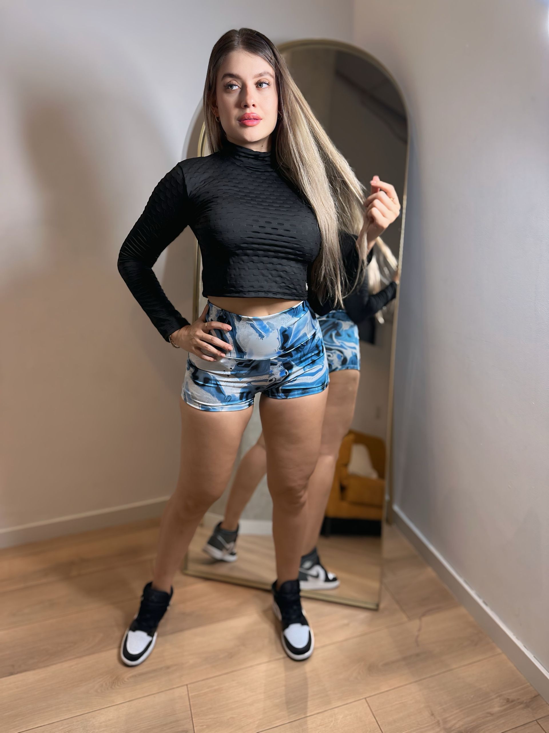 A woman in a black crop top and shorts is standing in front of a mirror.