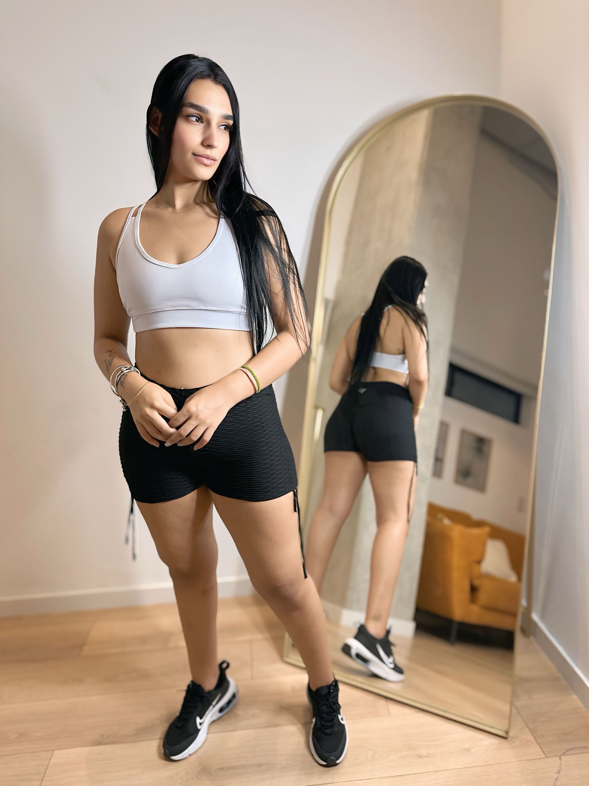 A woman in a white crop top and black shorts is standing in front of a mirror.