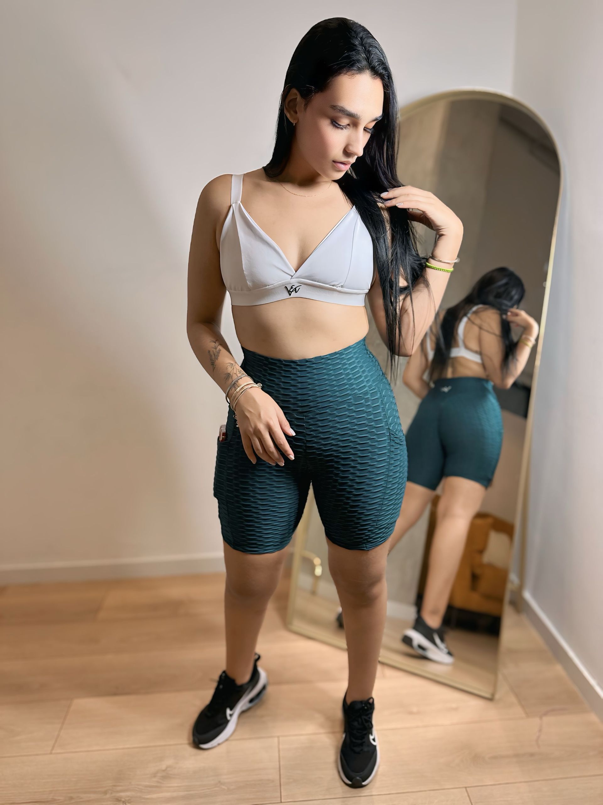 A woman in a white bra and green shorts is standing in front of a mirror.