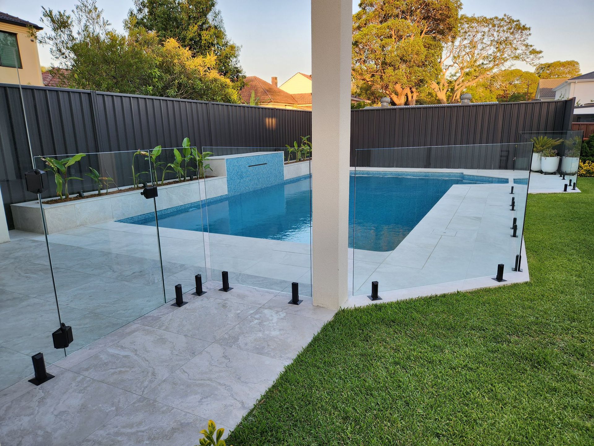 Backyard pool with glass safety fence, white patio, and green lawn under a covered patio — ClearView Glass Pool Fencing In Roselands, NSW