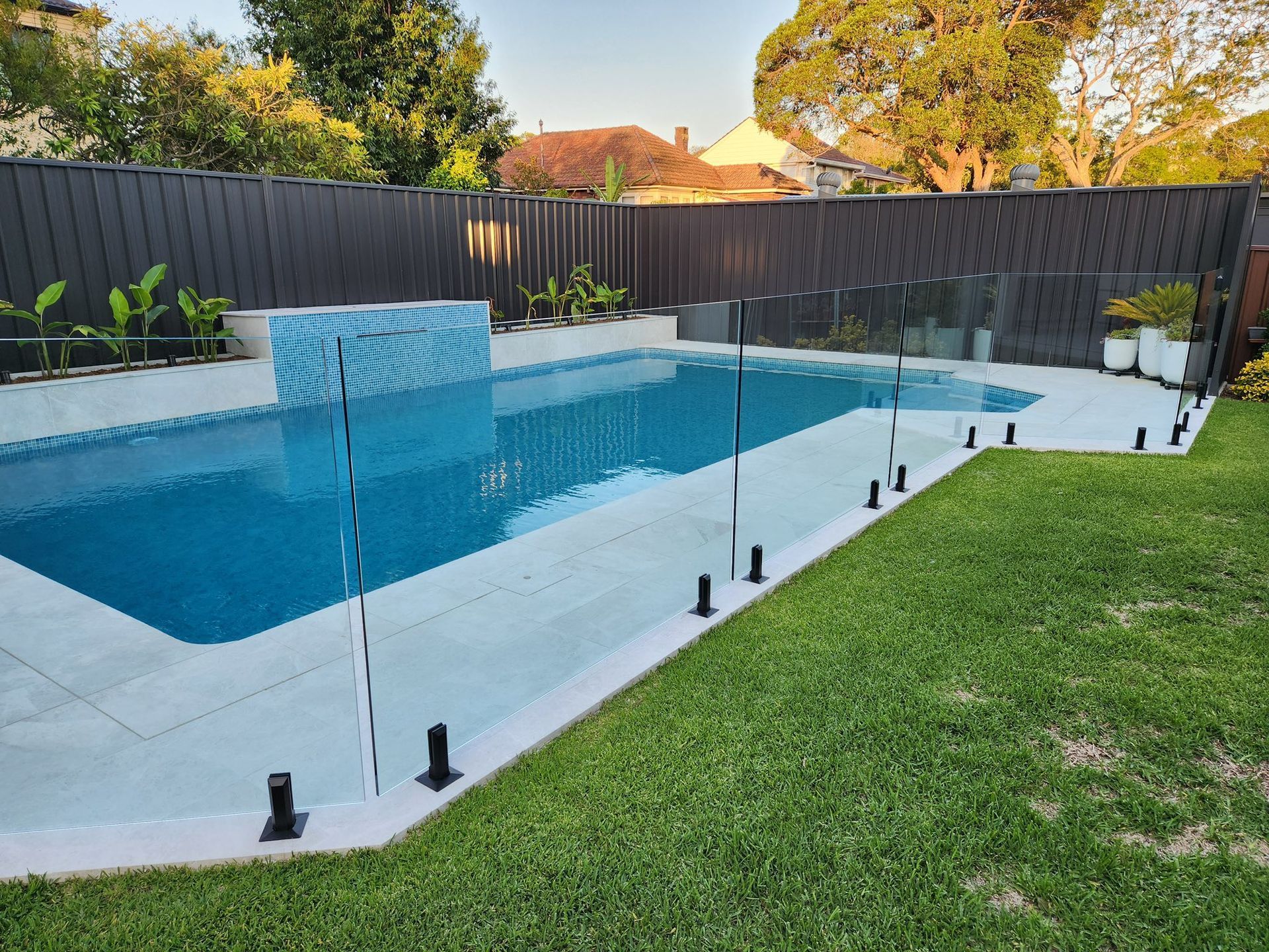 Modern backyard pool with glass fence, blue water, and green lawn at sunset — ClearView Glass Pool Fencing In Roselands, NSW