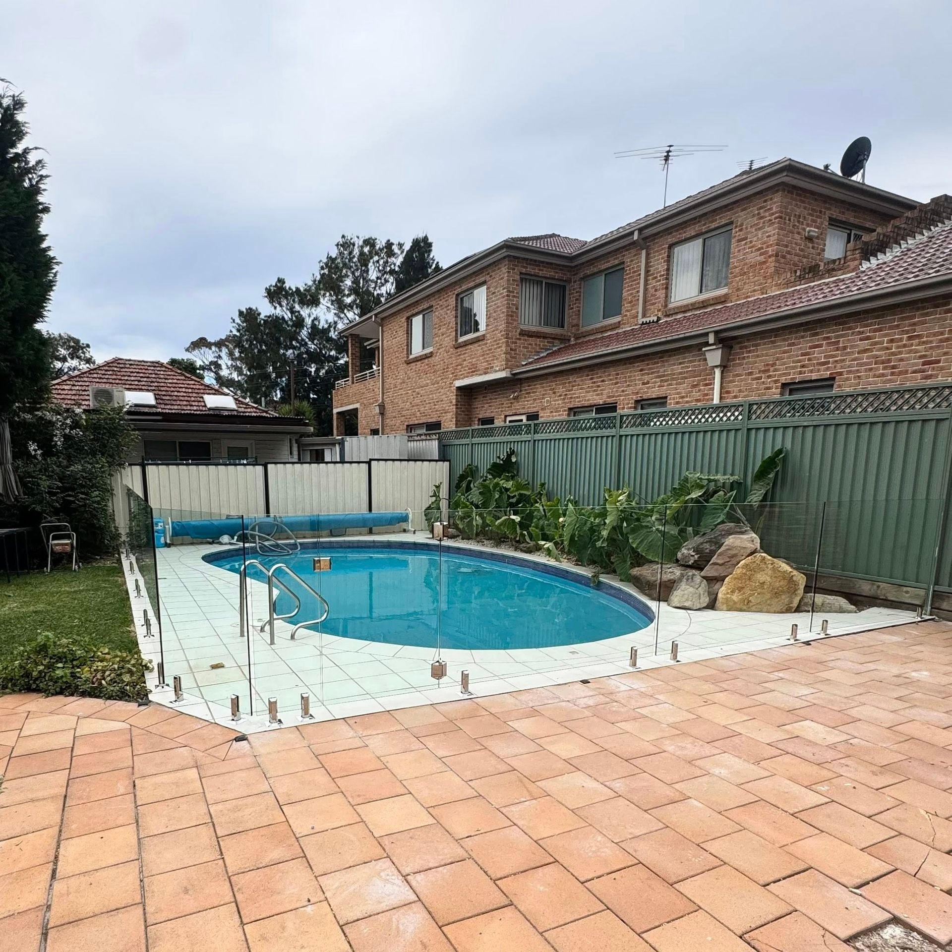 Backyard with a round swimming pool, brick patio, and two-story brick house behind a green fence — ClearView Glass Pool Fencing In Roselands, NSW
