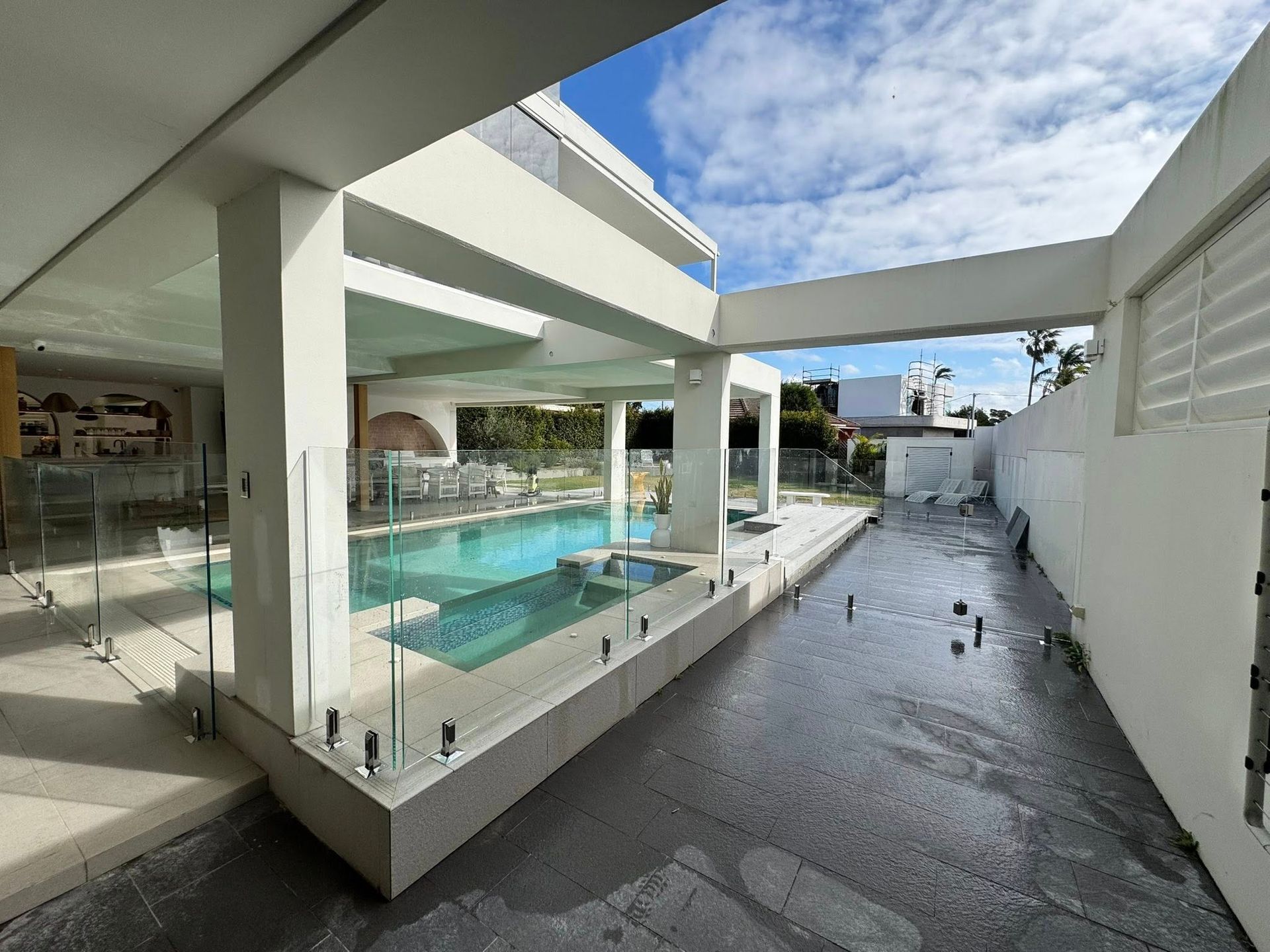 Modern courtyard with a turquoise pool, glass railing, white buildings, and a wet walkway under a partly cloudy sky — ClearView Glass Pool Fencing In Roselands, NSW