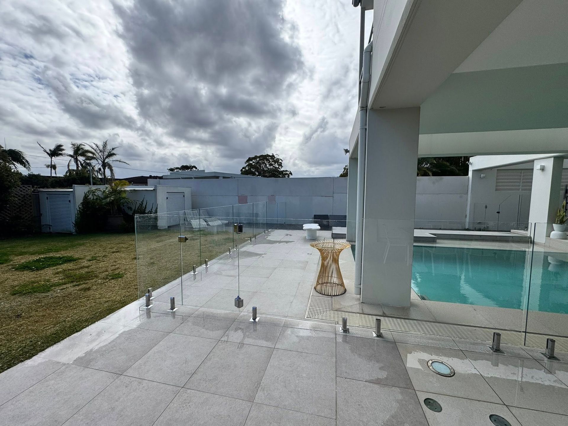 Back patio with pool, wet stone tiles, and cloudy sky beside a white house — ClearView Glass Pool Fencing In Roselands, NSW