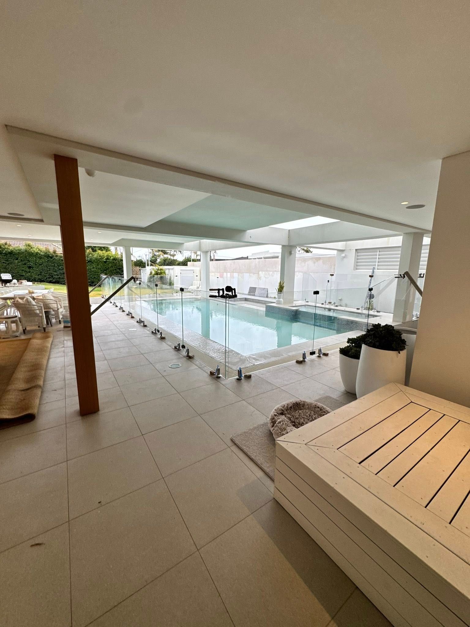 Covered poolside patio with lounge chairs, tiled floor, and a rectangular swimming pool under a canopy — ClearView Glass Pool Fencing In Roselands, NSW