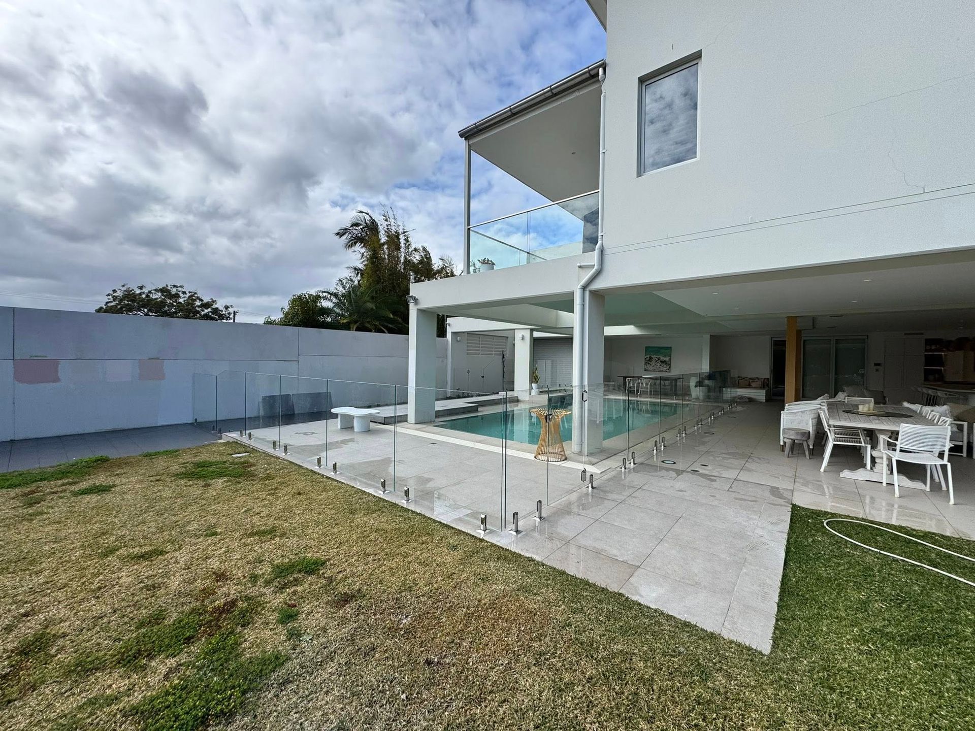 Modern white house with patio, pool, glass railing, and outdoor dining area under a cloudy sky — ClearView Glass Pool Fencing In Roselands, NSW