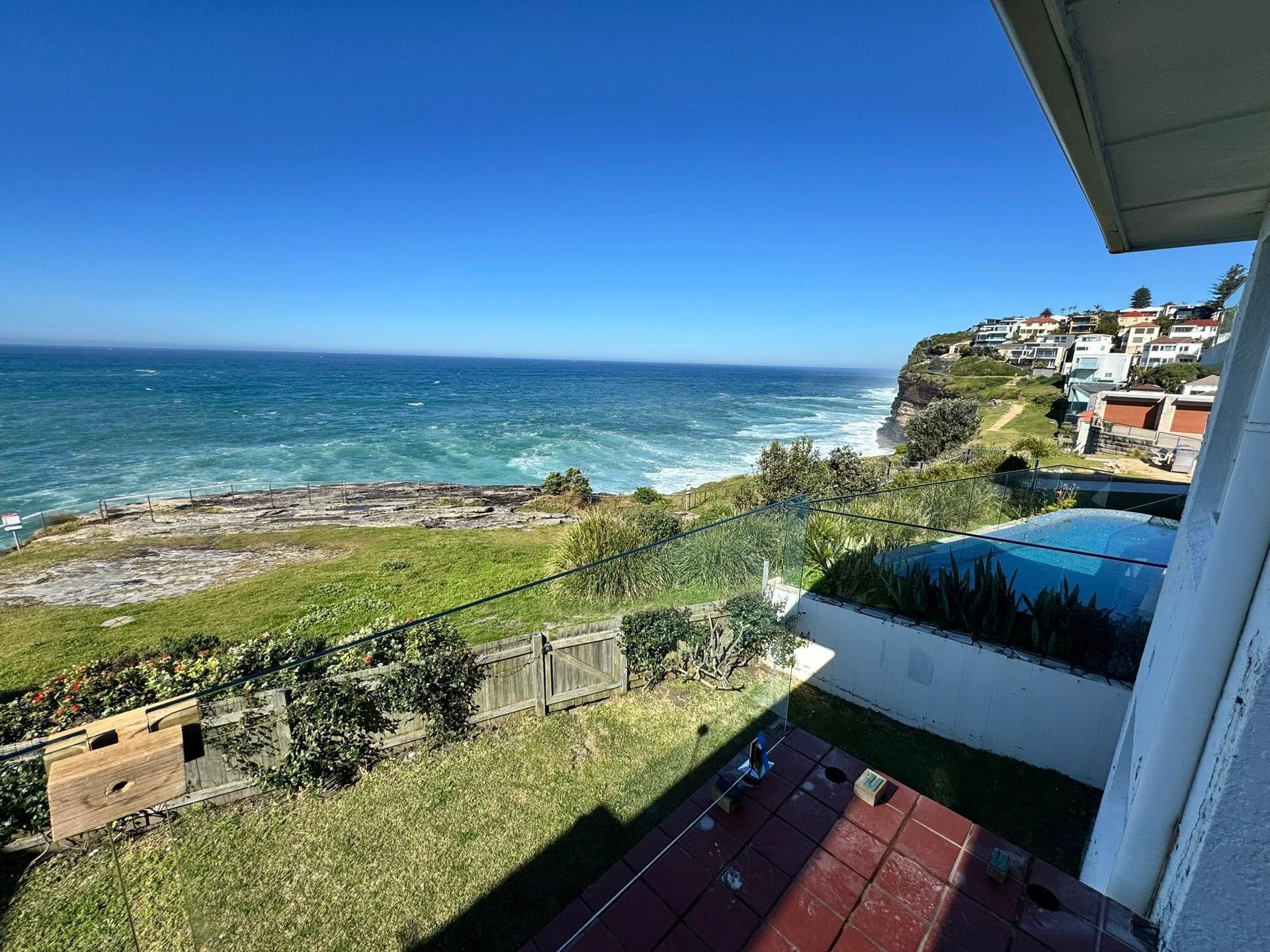 Ocean view from a balcony, with grassy cliffside, rocky shore, and houses under a clear blue sky. — ClearView Glass Pool Fencing In Northern Beaches, NSW