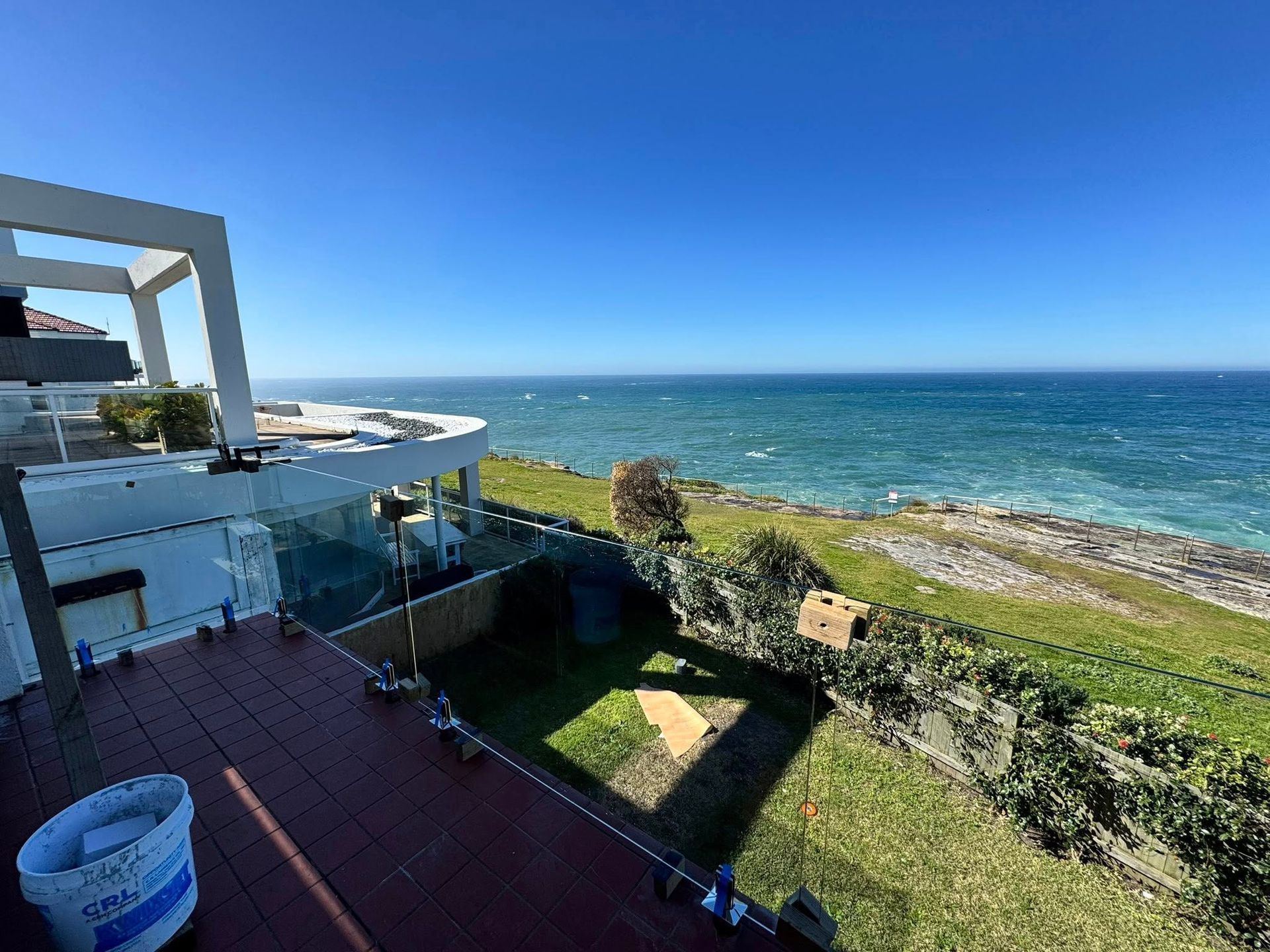 Oceanfront deck with glass railing, green lawn, and blue sea under a clear sky — ClearView Glass Pool Fencing In Roselands, NSW