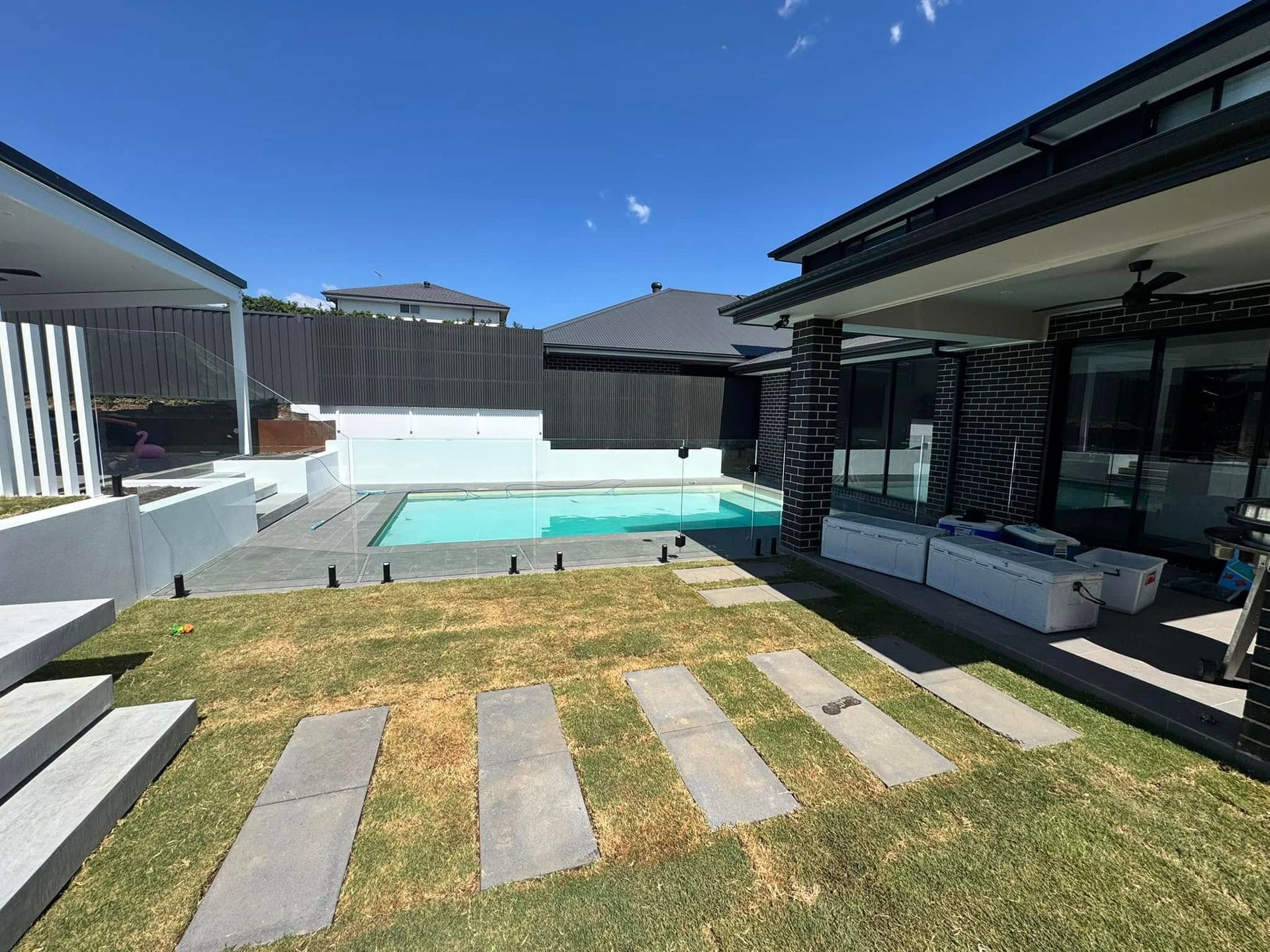 Backyard patio with pool, lawn, and modern house under a clear blue sky — ClearView Glass Pool Fencing In Roselands, NSW