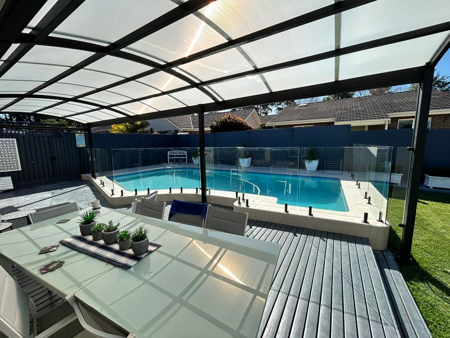 Covered outdoor pool with lounge chairs, benches, and potted plants under a black-framed canopy — ClearView Glass Pool Fencing In Roselands, NSW