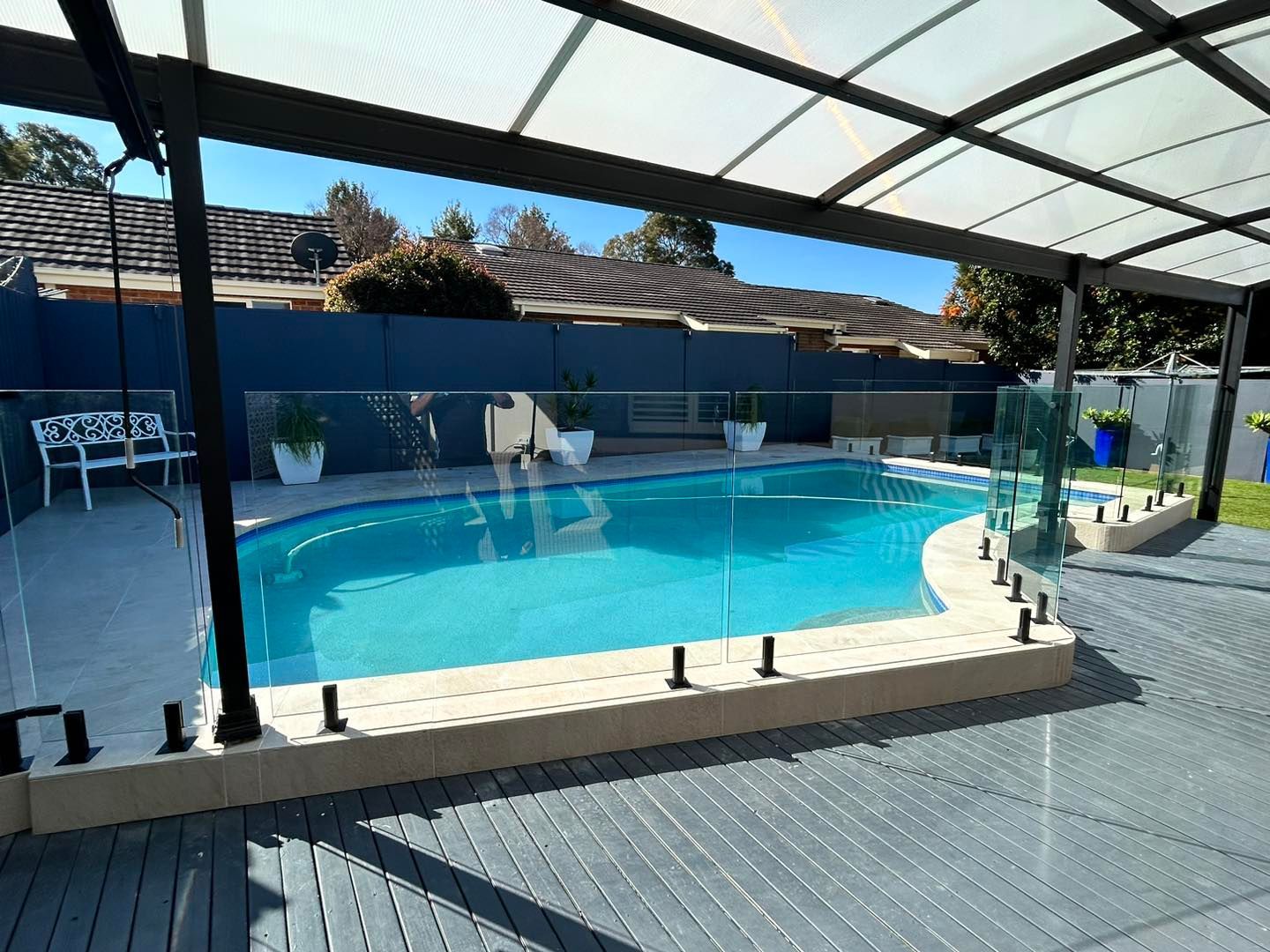 Covered backyard swimming pool with turquoise water, black fence, and patio decking under a translucent roof — ClearView Glass Pool Fencing In Roselands, NSW