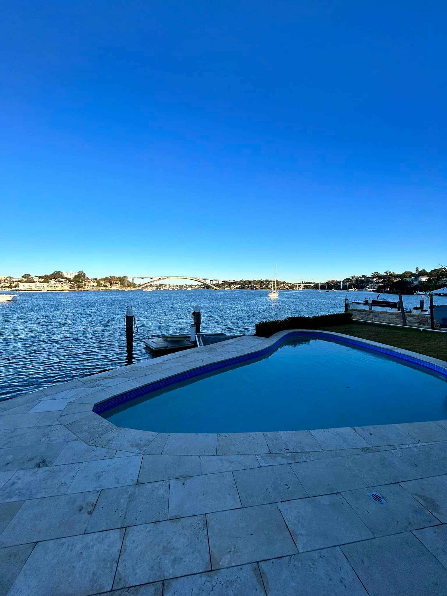 Blue swimming pool beside a waterfront patio at sunset, with calm water and distant shoreline — ClearView Glass Pool Fencing In Roselands, NSW