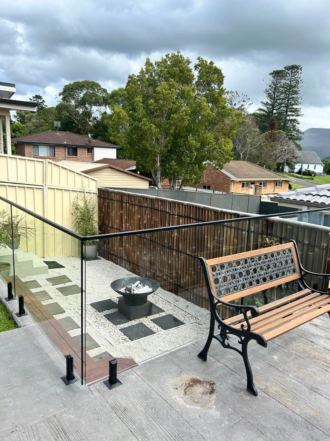 Backyard patio with bench, tiled floor, glass railing, and distant trees and houses — ClearView Glass Pool Fencing In Sutherland Shire, NSW