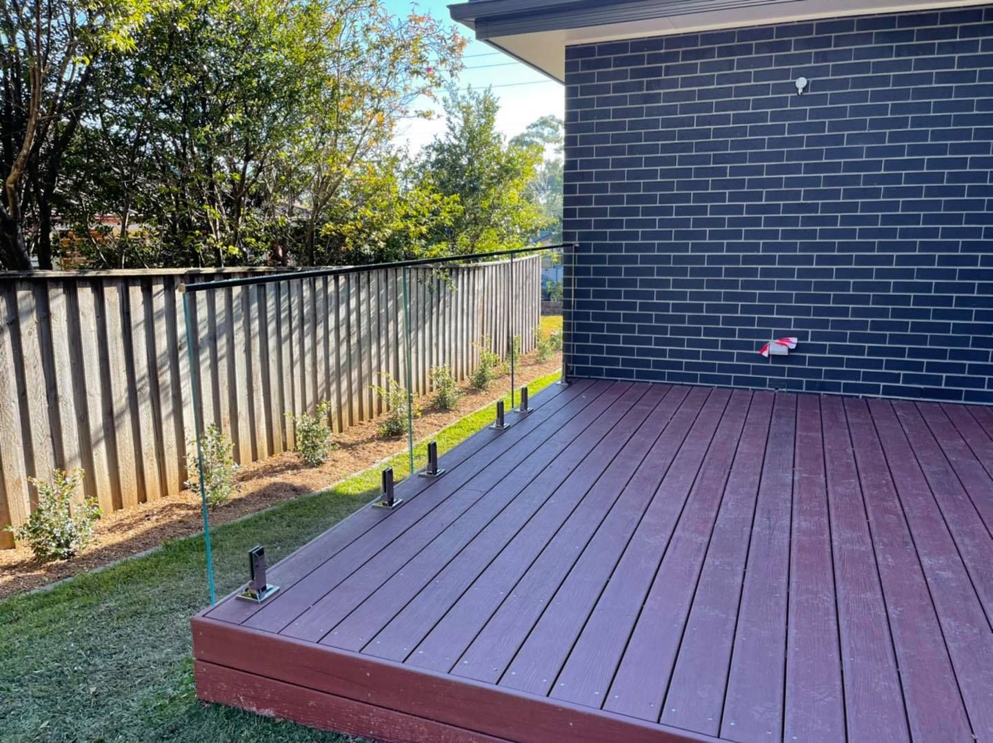 Backyard deck with gray fence, dark brick wall, and a red-brown wooden platform under a clear sky — ClearView Glass Pool Fencing In Roselands, NSW