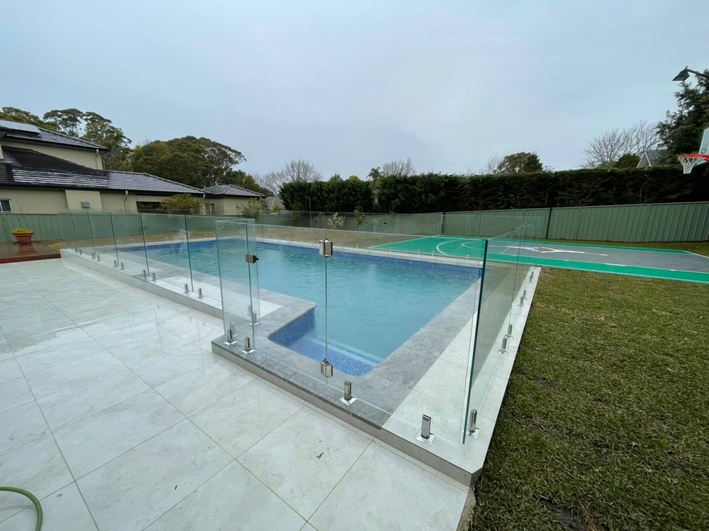 Outdoor swimming pool with clear water, glass safety fence, and tiled deck beside a grassy lawn — ClearView Glass Pool Fencing In Roselands, NSW