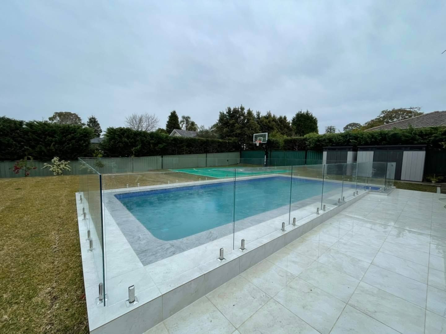 Outdoor swimming pool with glass fence and tiled deck under a cloudy sky — ClearView Glass Pool Fencing In Roselands, NSW