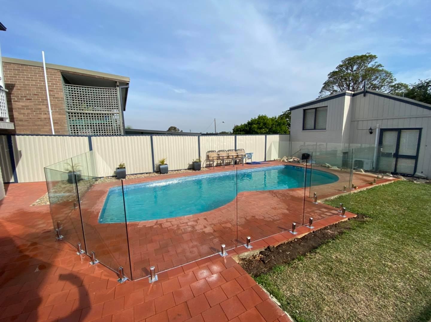 Backyard swimming pool surrounded by red brick patio and safety fence under a cloudy sky — ClearView Glass Pool Fencing In Roselands, NSW