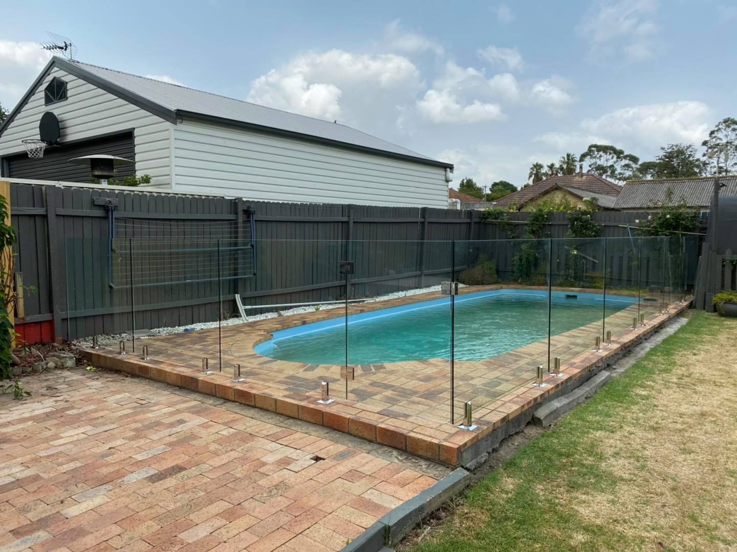 Backyard with a fenced in-ground pool, brick patio, and grass lawn — ClearView Glass Pool Fencing In Roselands, NSW