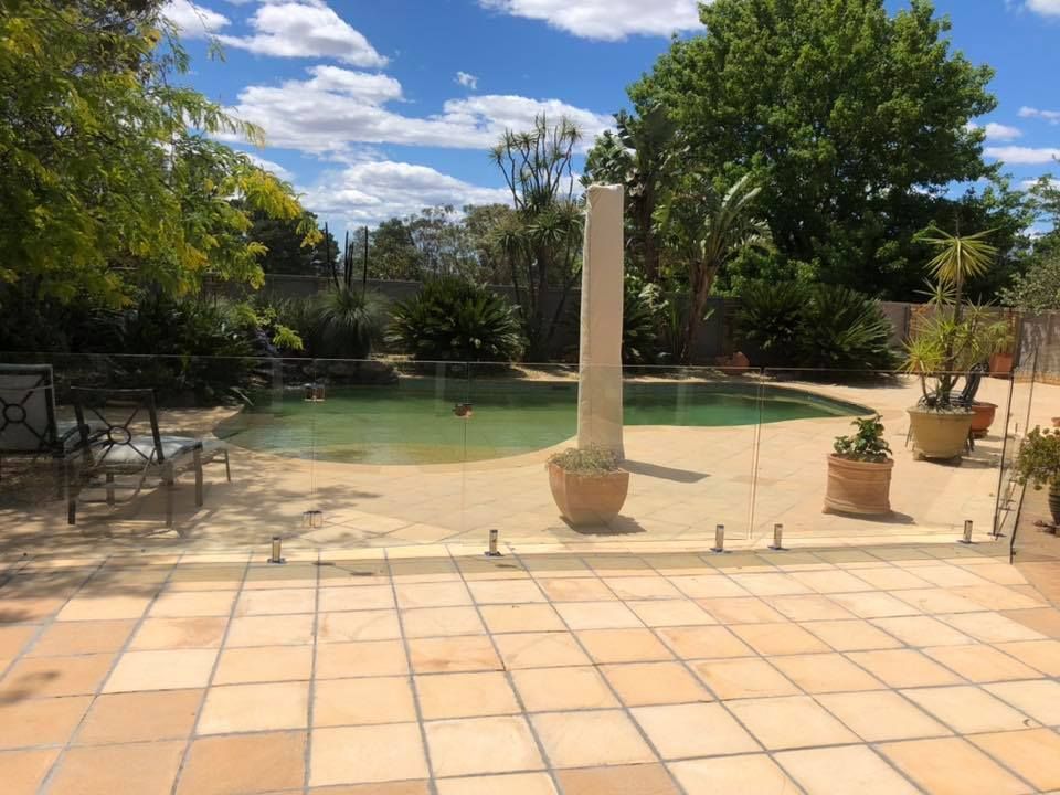 Sunlit backyard patio with terracotta tiles, a pool, potted plants, and trees under a blue sky — ClearView Glass Pool Fencing In Roselands, NSW