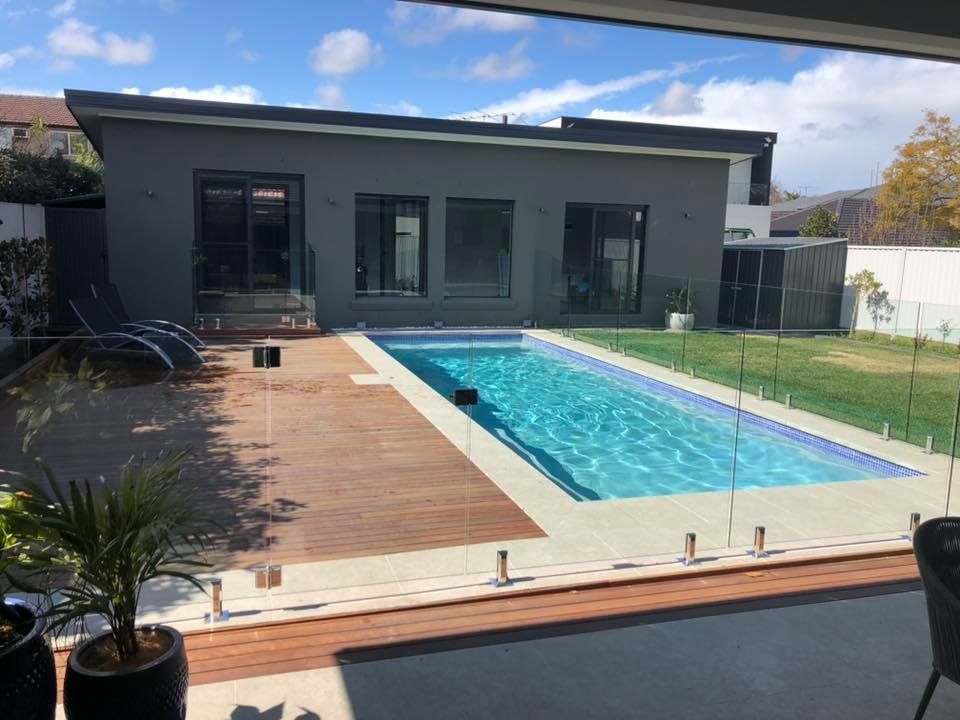 Backyard patio with a long rectangular swimming pool beside a modern gray house and wooden deck — ClearView Glass Pool Fencing In Roselands, NSW