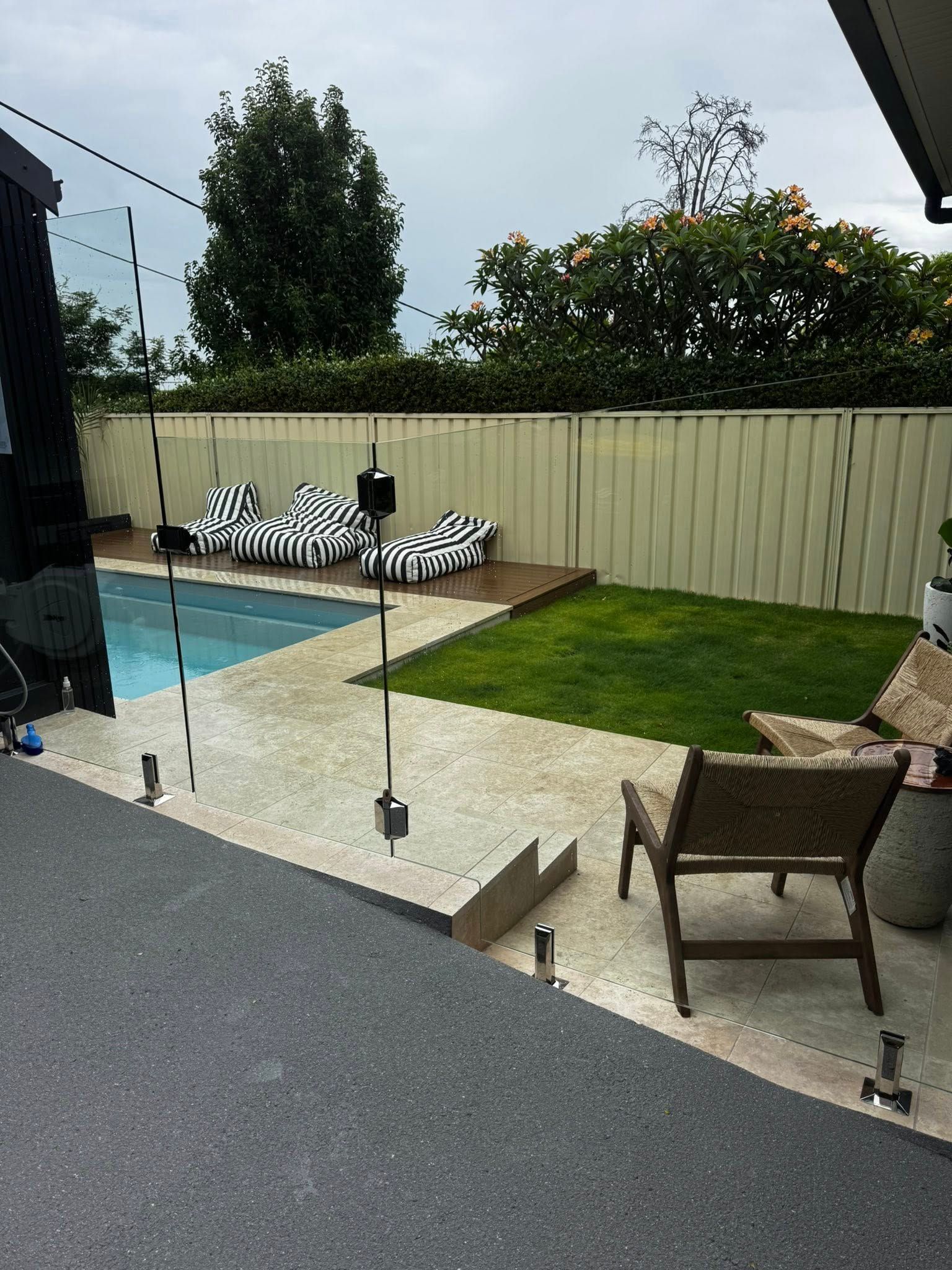 Backyard pool patio with striped loungers, lawn, and chairs beside a glass fence — ClearView Glass Pool Fencing In Roselands, NSW