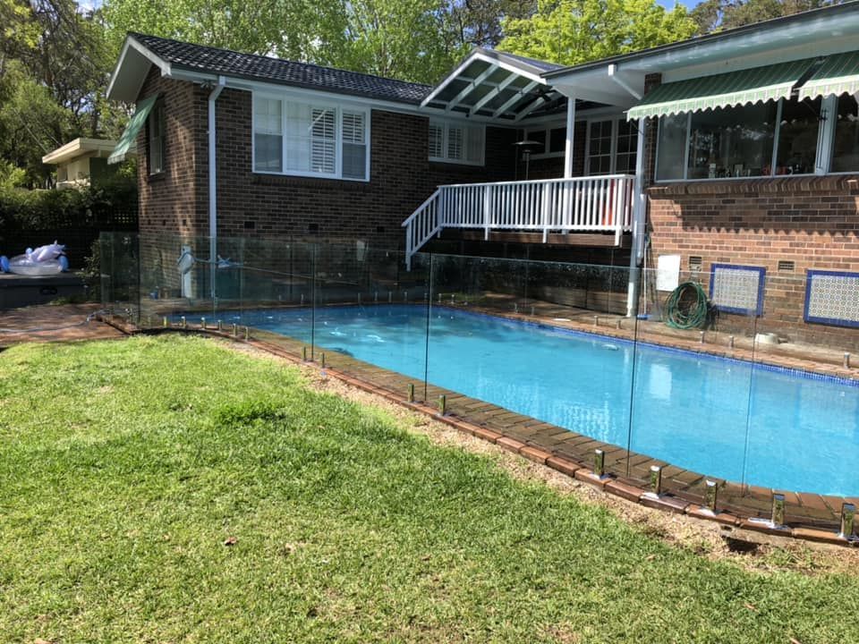 Backyard swimming pool beside a brick house and screened patio, with green lawn in front — ClearView Glass Pool Fencing In Roselands, NSW