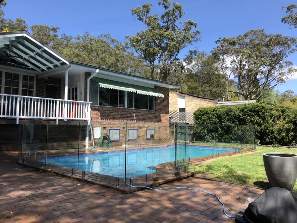 Backyard pool beside a brick house with a covered patio and greenery under a blue sky — ClearView Glass Pool Fencing In Roselands, NSW