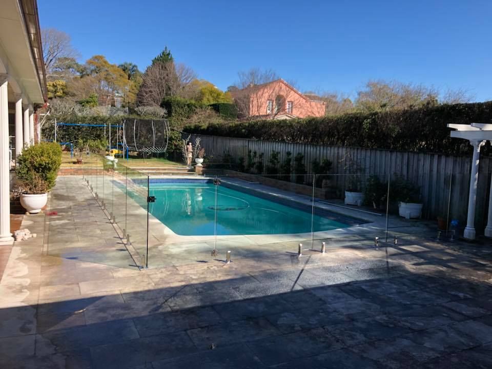 Backyard swimming pool with glass fence, tiled patio, and hedges under a clear blue sky — ClearView Glass Pool Fencing In Roselands, NSW