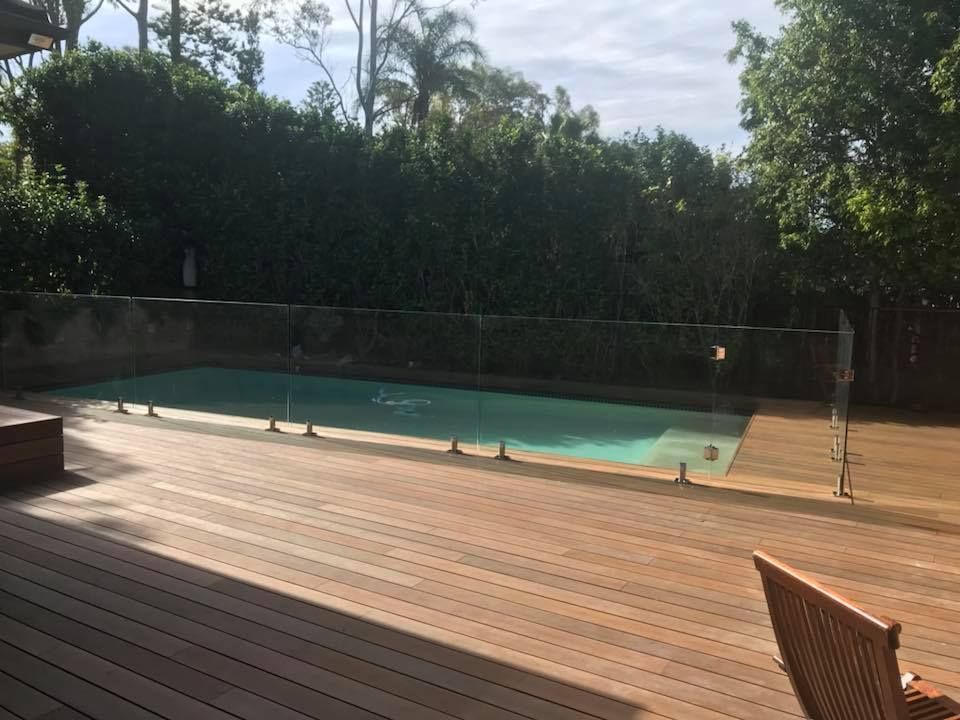 Backyard pool beside a wooden deck, with trees and a clear sky in the background — ClearView Glass Pool Fencing In Roselands, NSW