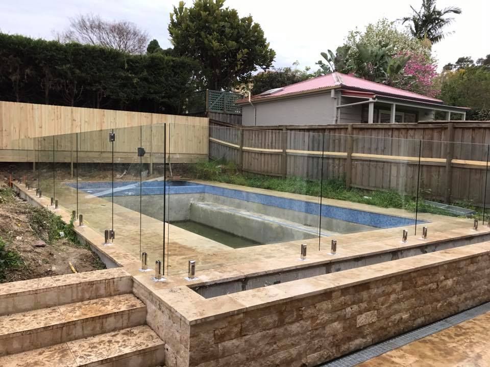 Empty backyard swimming pool under construction, surrounded by glass fencing and a wooden fence — ClearView Glass Pool Fencing In Roselands, NSW