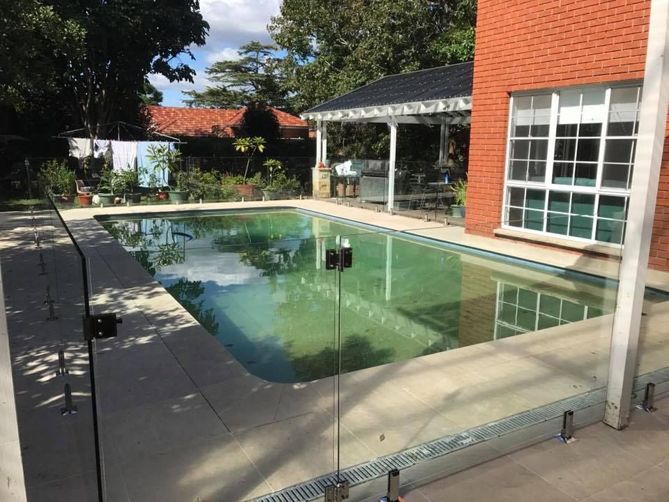 Backyard swimming pool beside a brick house and covered patio, with lounge chairs and trees reflected in water — ClearView Glass Pool Fencing In Roselands, NSW