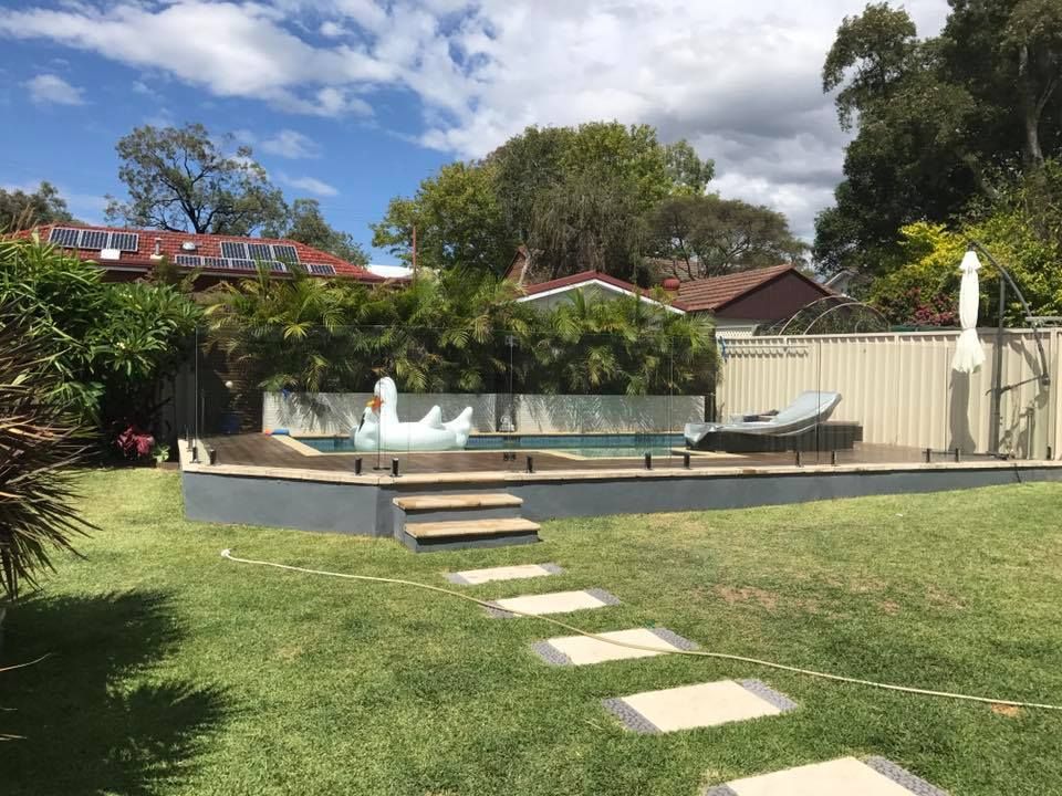 Backyard pool with stone path, lawn, white fence, and patio seating under a partly cloudy sky — ClearView Glass Pool Fencing In Roselands, NSW