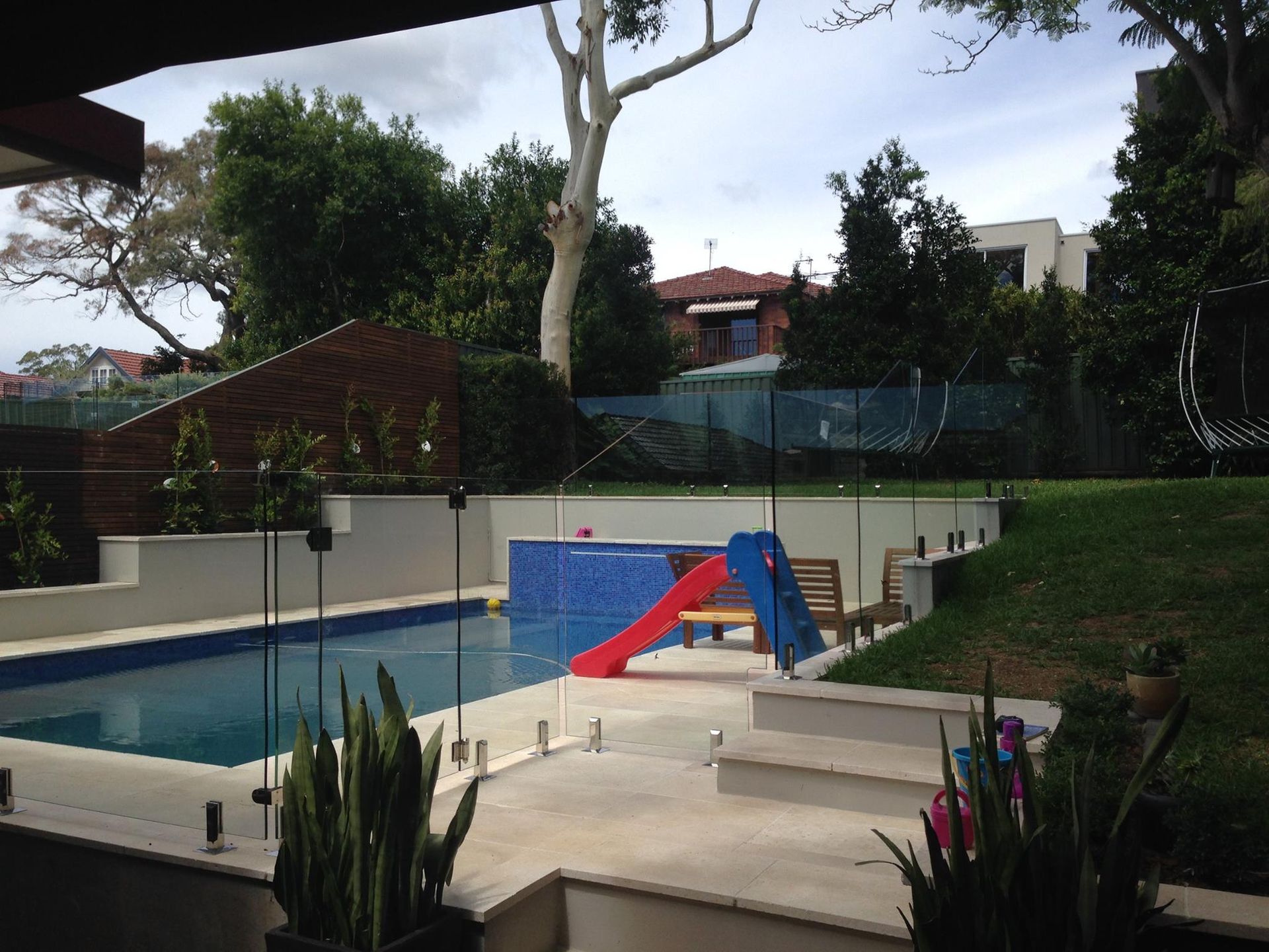 Backyard swimming pool with a red slide, blue water, and surrounding patio and greenery — ClearView Glass Pool Fencing In Roselands, NSW