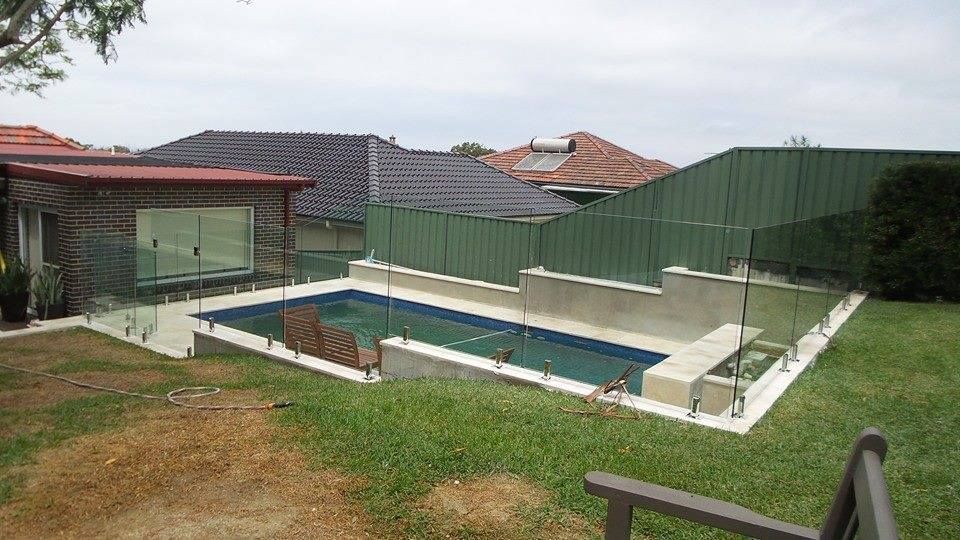 Backyard with a rectangular in-ground pool, white pool deck, and green fencing beside houses — ClearView Glass Pool Fencing In Roselands, NSW