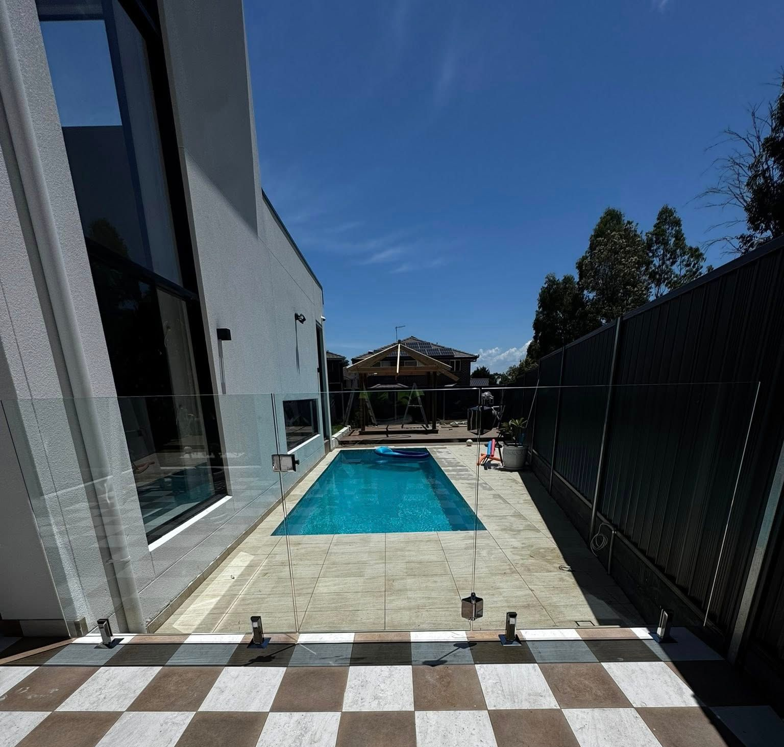 Backyard patio with a rectangular blue pool, modern house, and black fence under a clear sky — ClearView Glass Pool Fencing In Roselands, NSW
