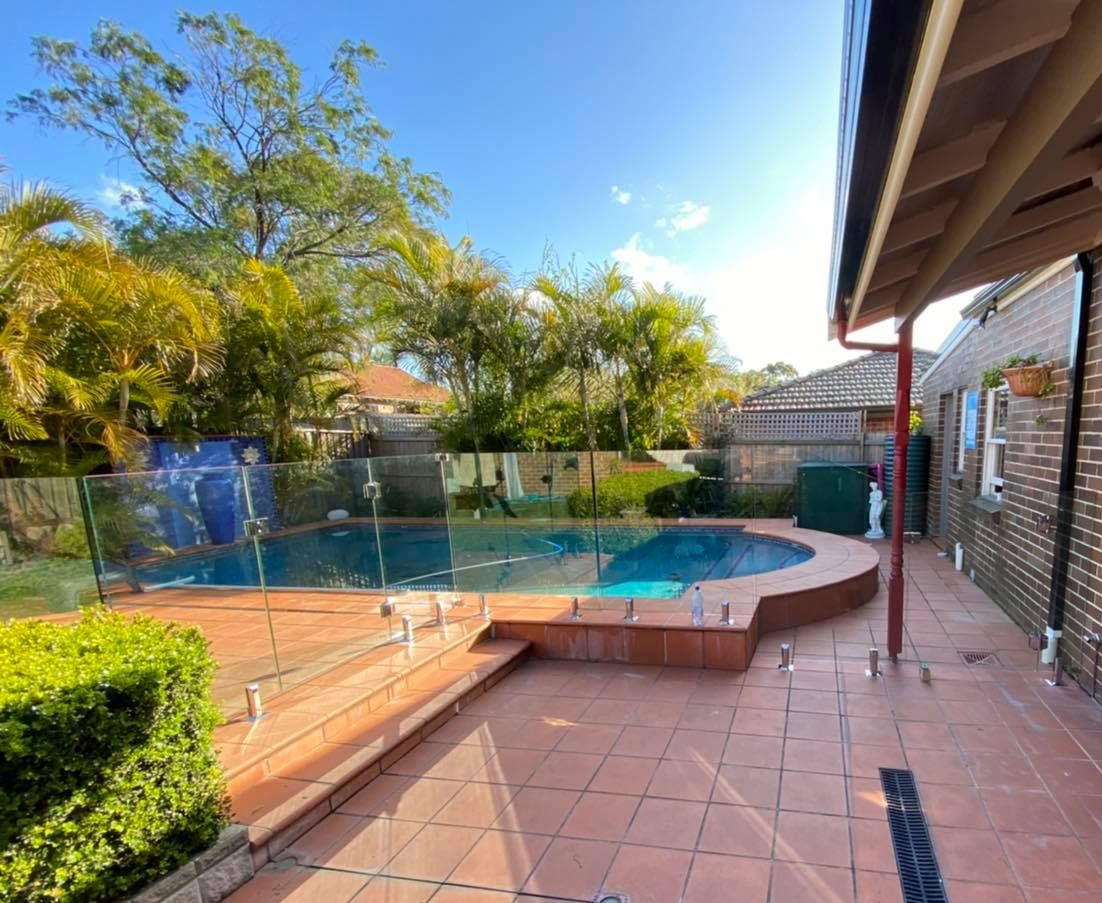 A Backyard Patio With a Curved Swimming Pool and a Glass Fence — ClearView Glass Pool Fencing In Eastern Suburbs, NSW
