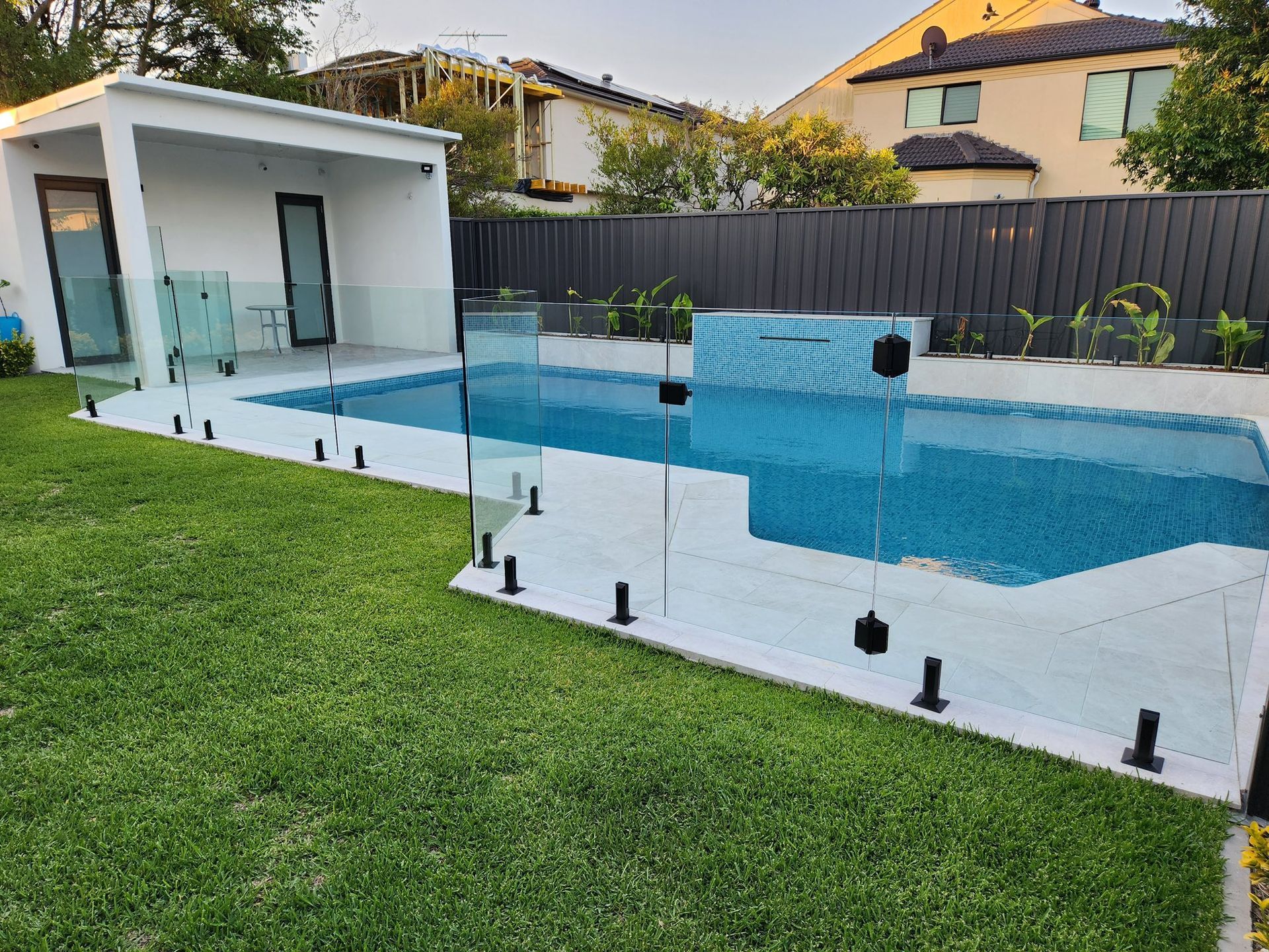 Backyard pool with white tile deck, green lawn, black fence, and a small pool house — ClearView Glass Pool Fencing In Roselands, NSW