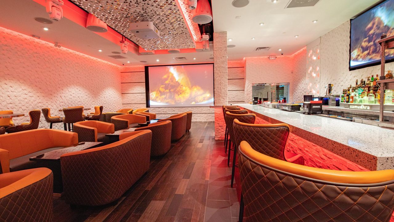 Interior of a modern bar with orange seating, a white bar, a large screen, and red lighting.
