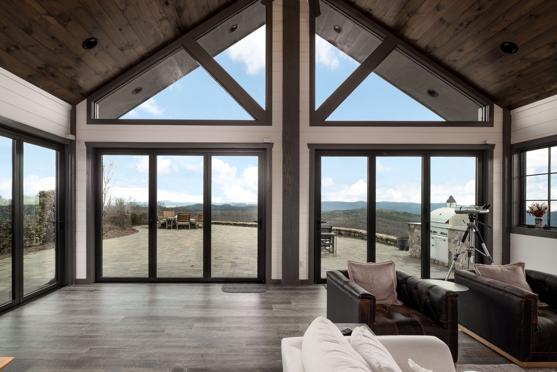 Interior of a cabin with large windows overlooking a mountain view, including a telescope and leather chairs.