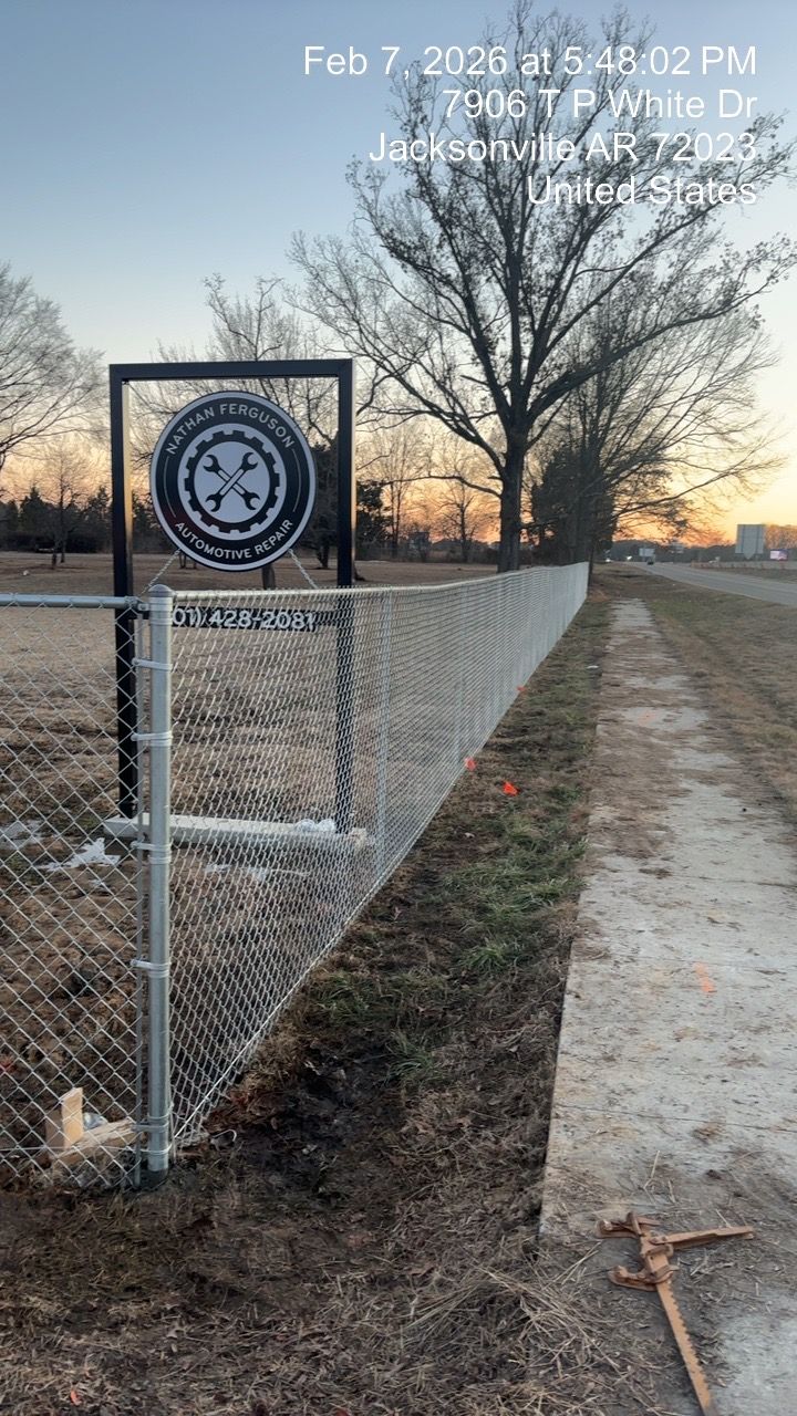 Chain-link fence with a sign beside a dirt path and leafless trees at sunset.