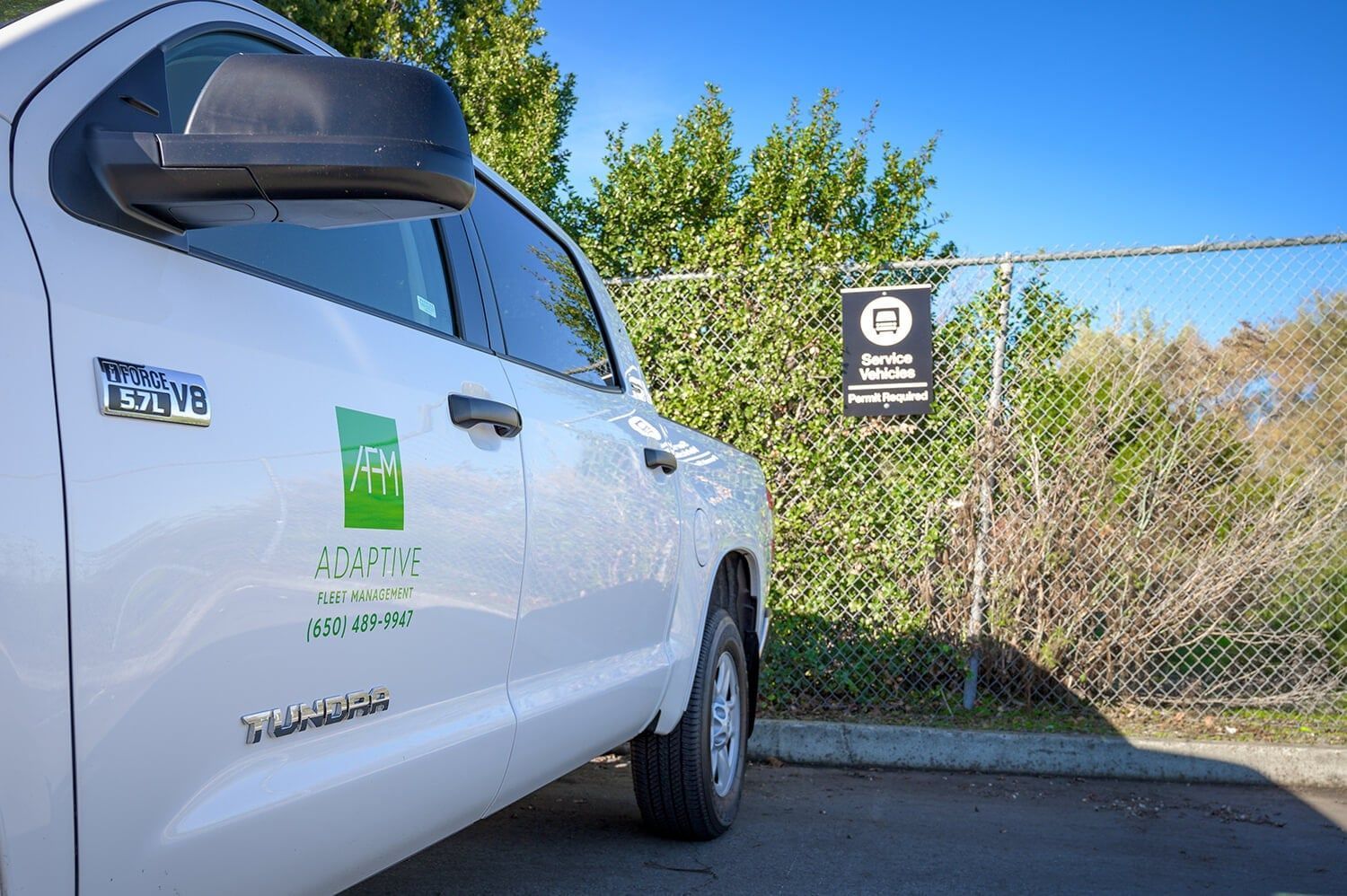 White utility truck parked by a chain-link fence with a green logo on the door. | Adaptive Fleet Management